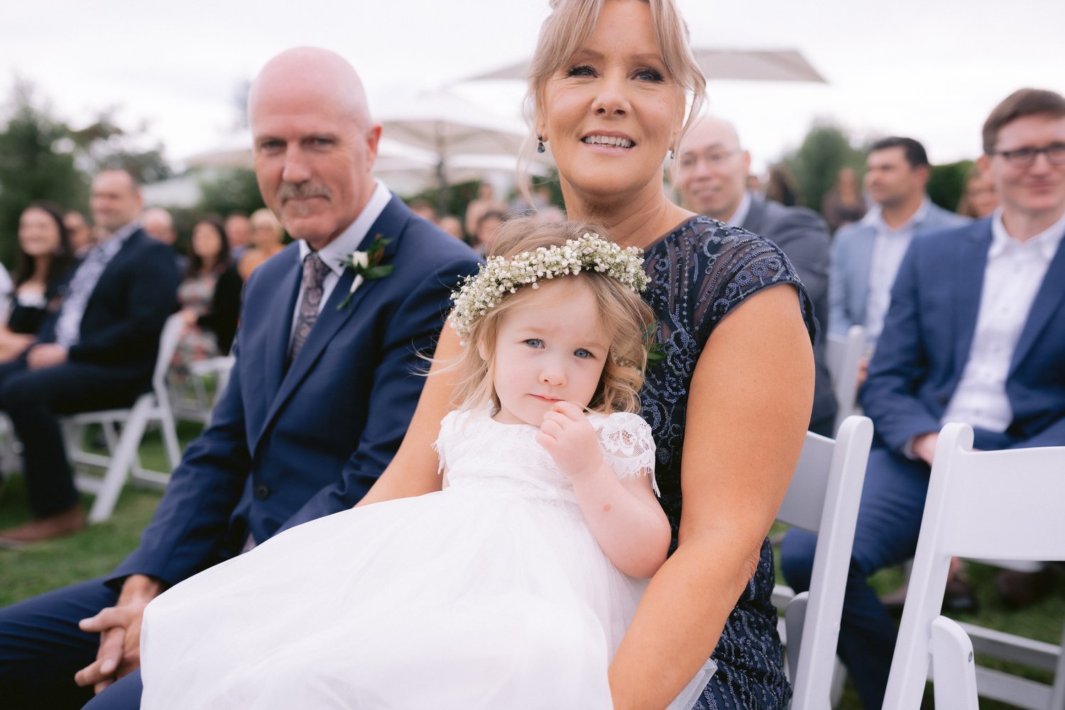 A woman holding a young girl in a white dress with a floral headband, seated outdoors at an event with other attendees in the background.