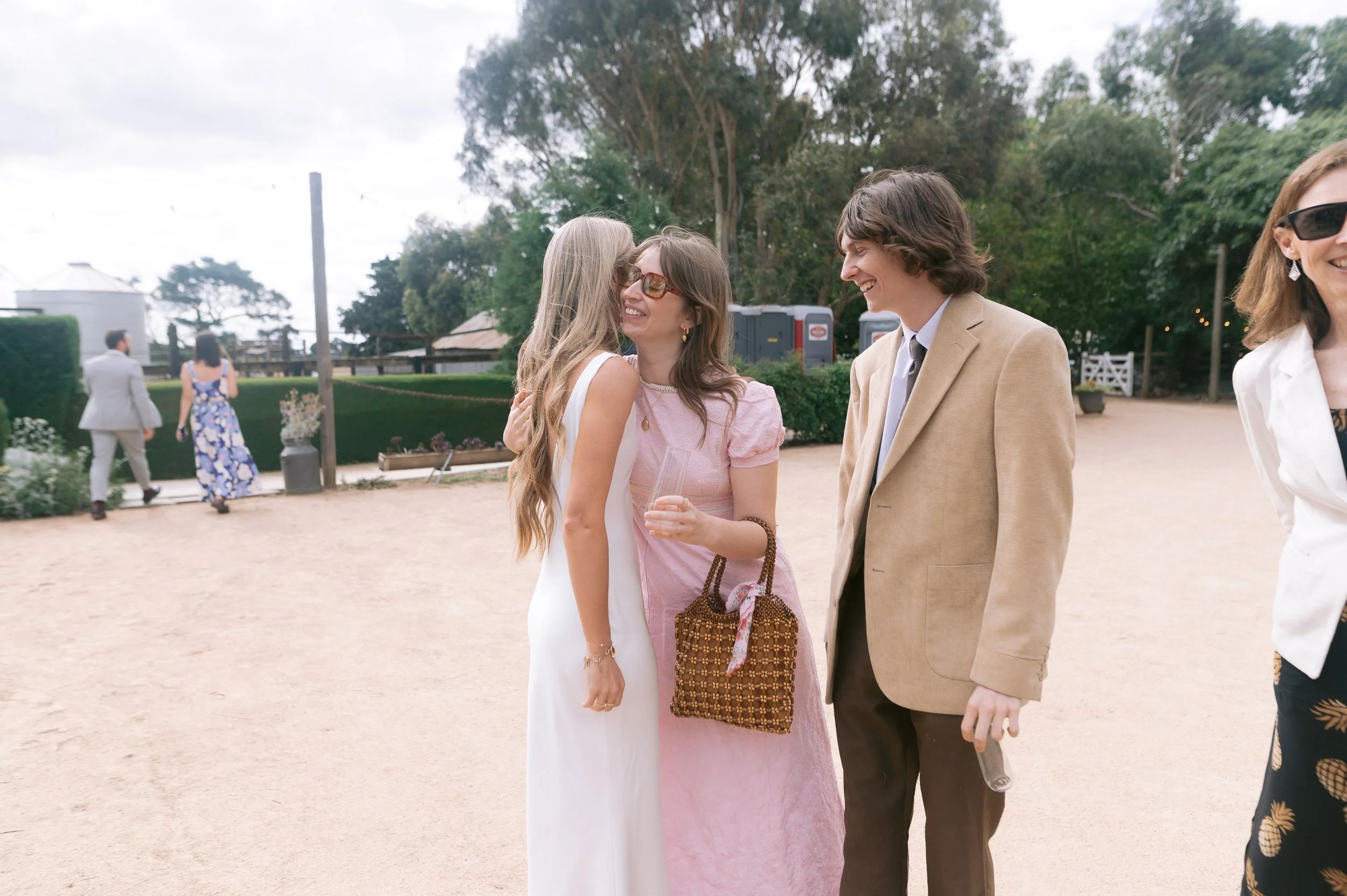 Three people smiling and hugging at an outdoor event, with two women and one man, dressed in semi-formal attire, on a sandy area with greenery and other guests in the background.