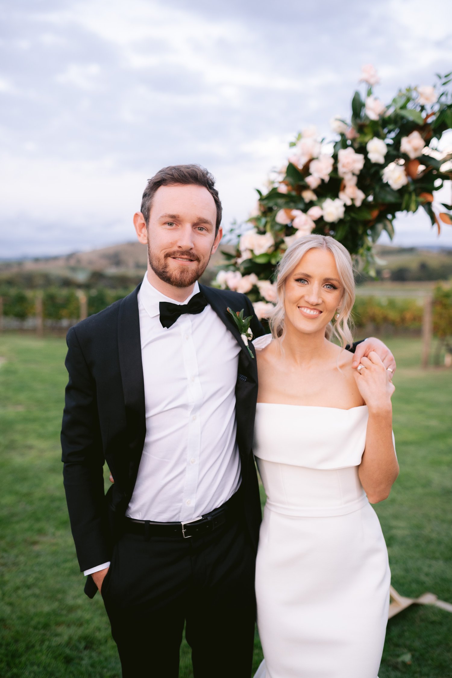 Bride and groom posing in formal attire outdoors with floral arch and scenic background.
