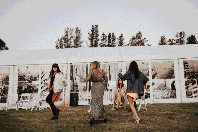 Women dancing outdoors on a lawn in front of a large white tent at a casual gathering.