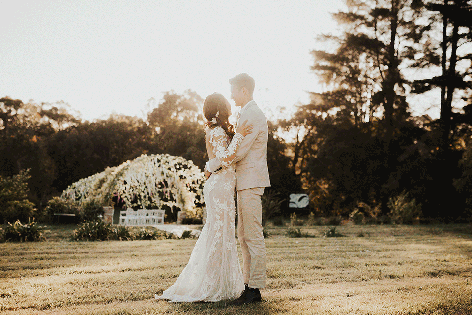 Bride and groom in wedding attire embracing outdoors during sunset, standing on a lawn with trees and floral arch in the background.