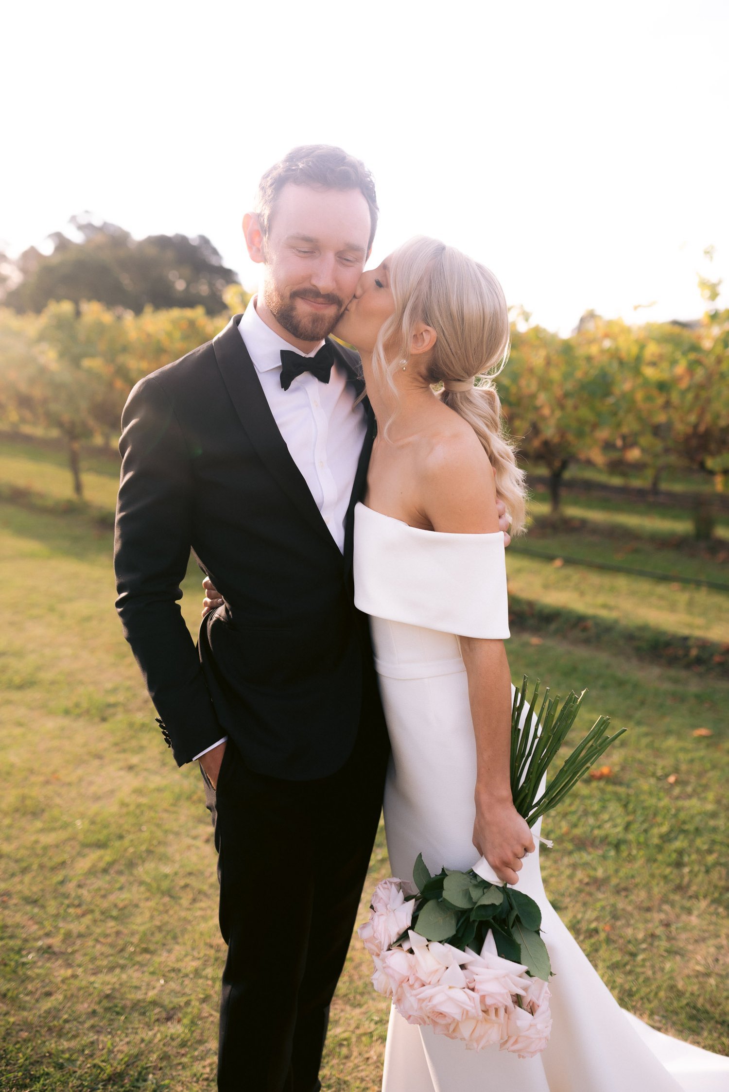 Bride kissing groom on the cheek, holding bouquet, outdoors in vineyard.