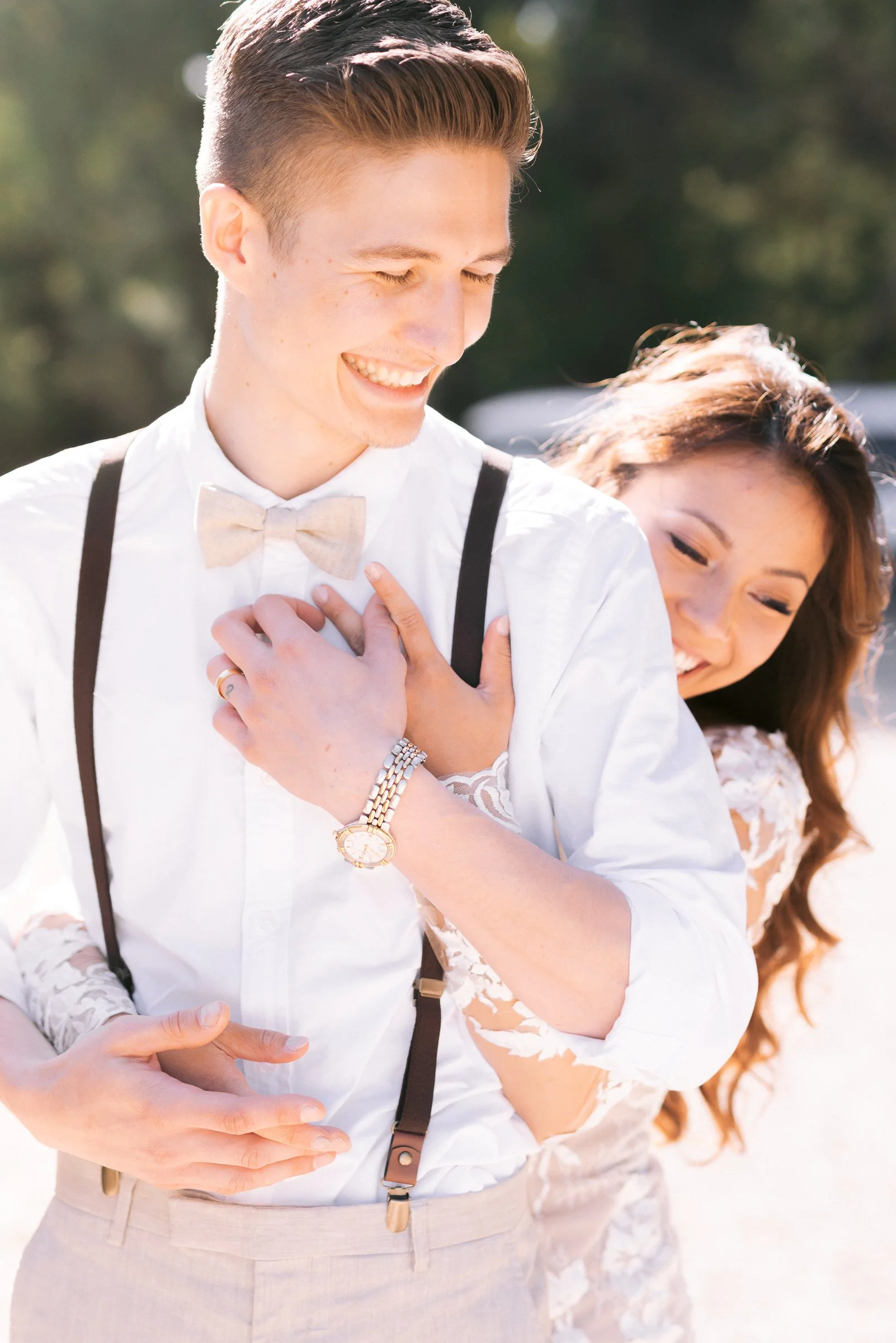 A couple embracing outdoors, woman in a lace dress, man wearing a bow tie and suspenders.