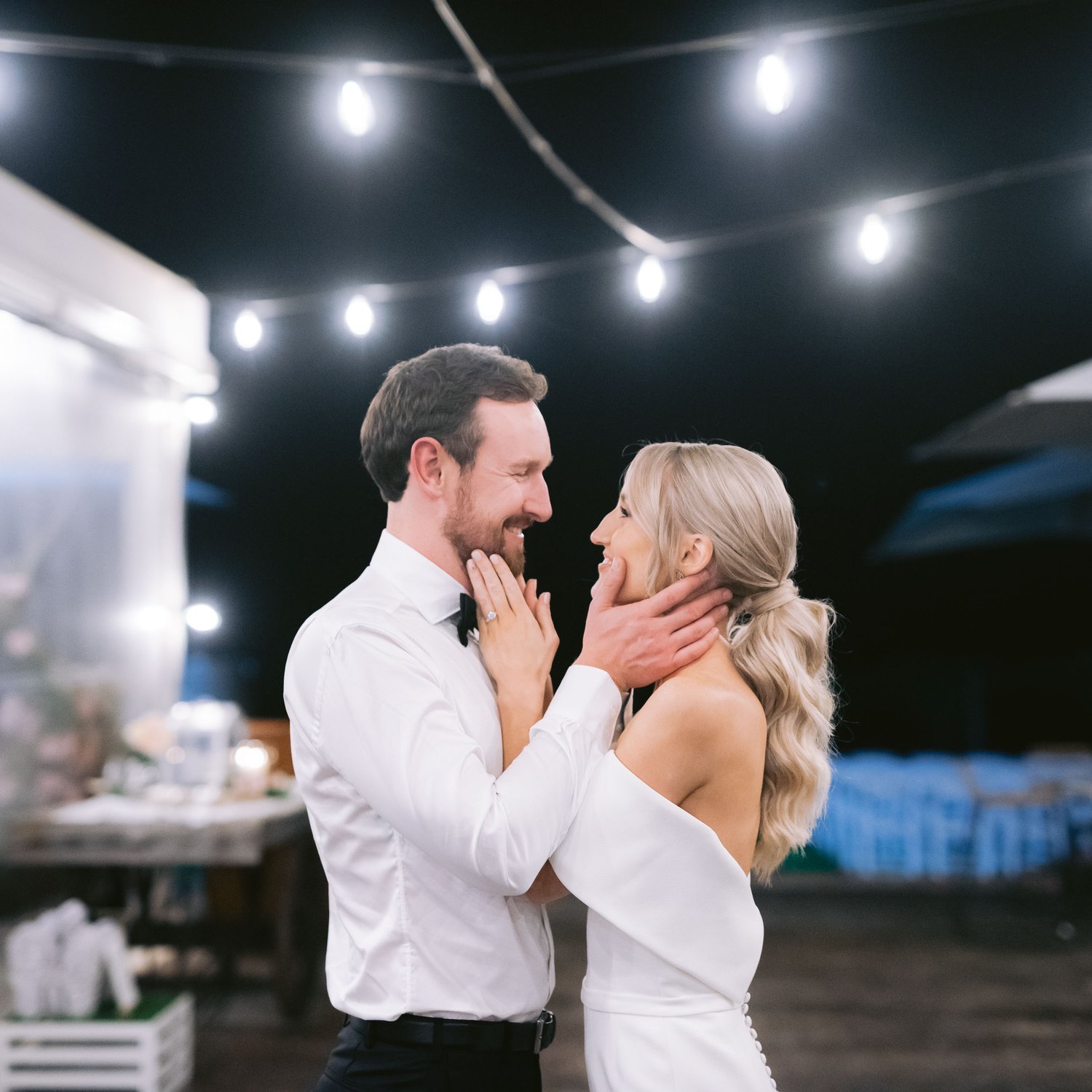 A couple in wedding attire embracing under string lights at night.