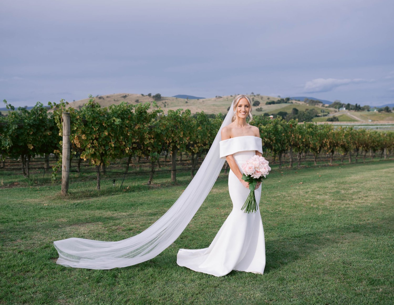 Bride in a wedding dress holding a bouquet of pink roses standing in a vineyard with hills in the background.