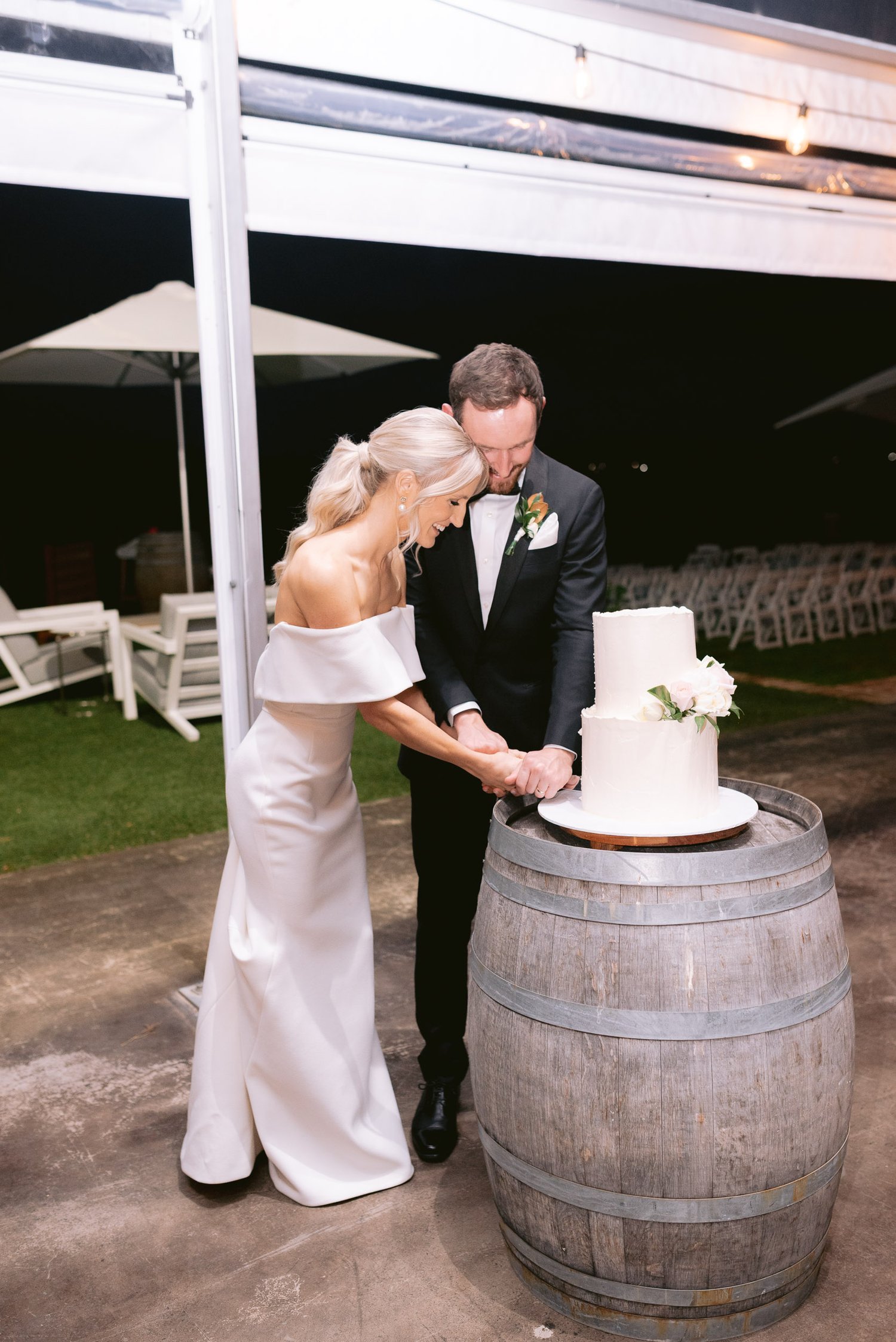 Bride and groom cutting a wedding cake on a wooden barrel at an outdoor venue.