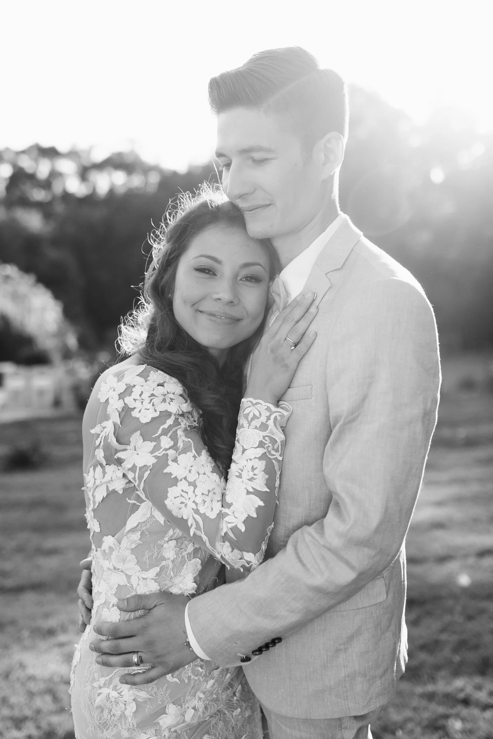 Black and white photo of a smiling couple embracing outdoors, wearing formal attire, with a blurred natural background.