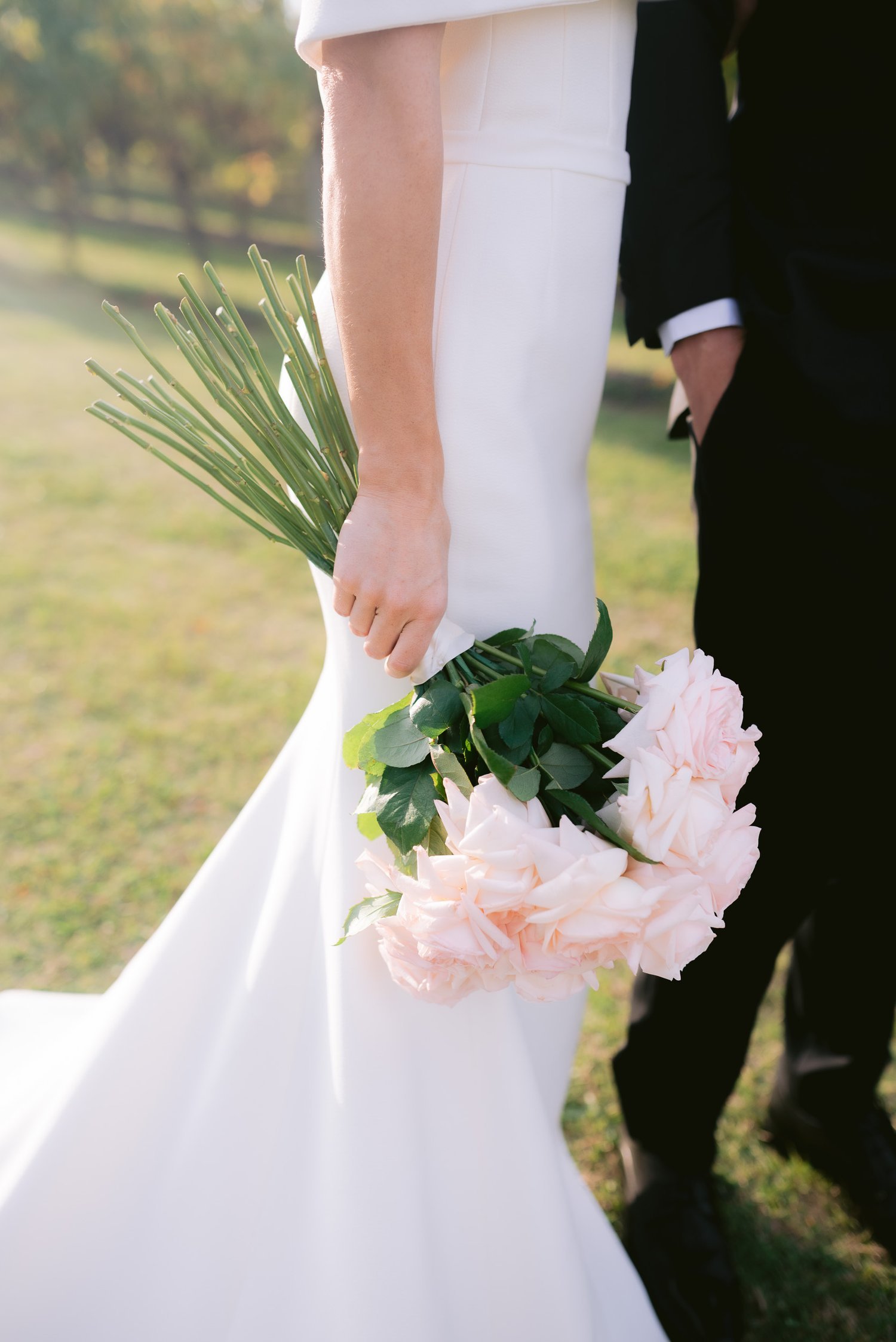 Bride holding a bouquet of light pink roses, wearing a white wedding dress, standing outside on grass with a person in a black suit.