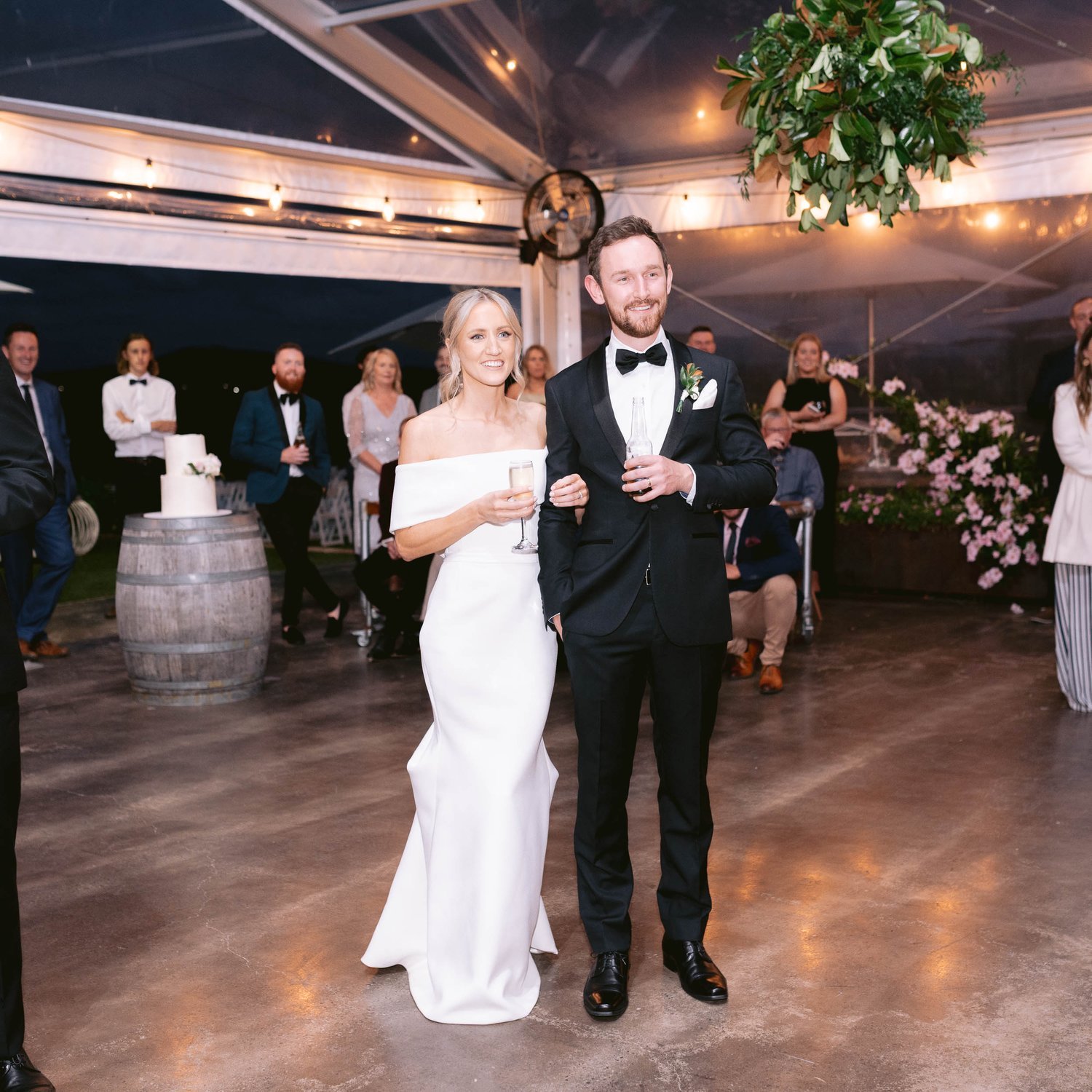 Bride and groom holding glasses, smiling at their wedding reception with guests and a cake in the background.