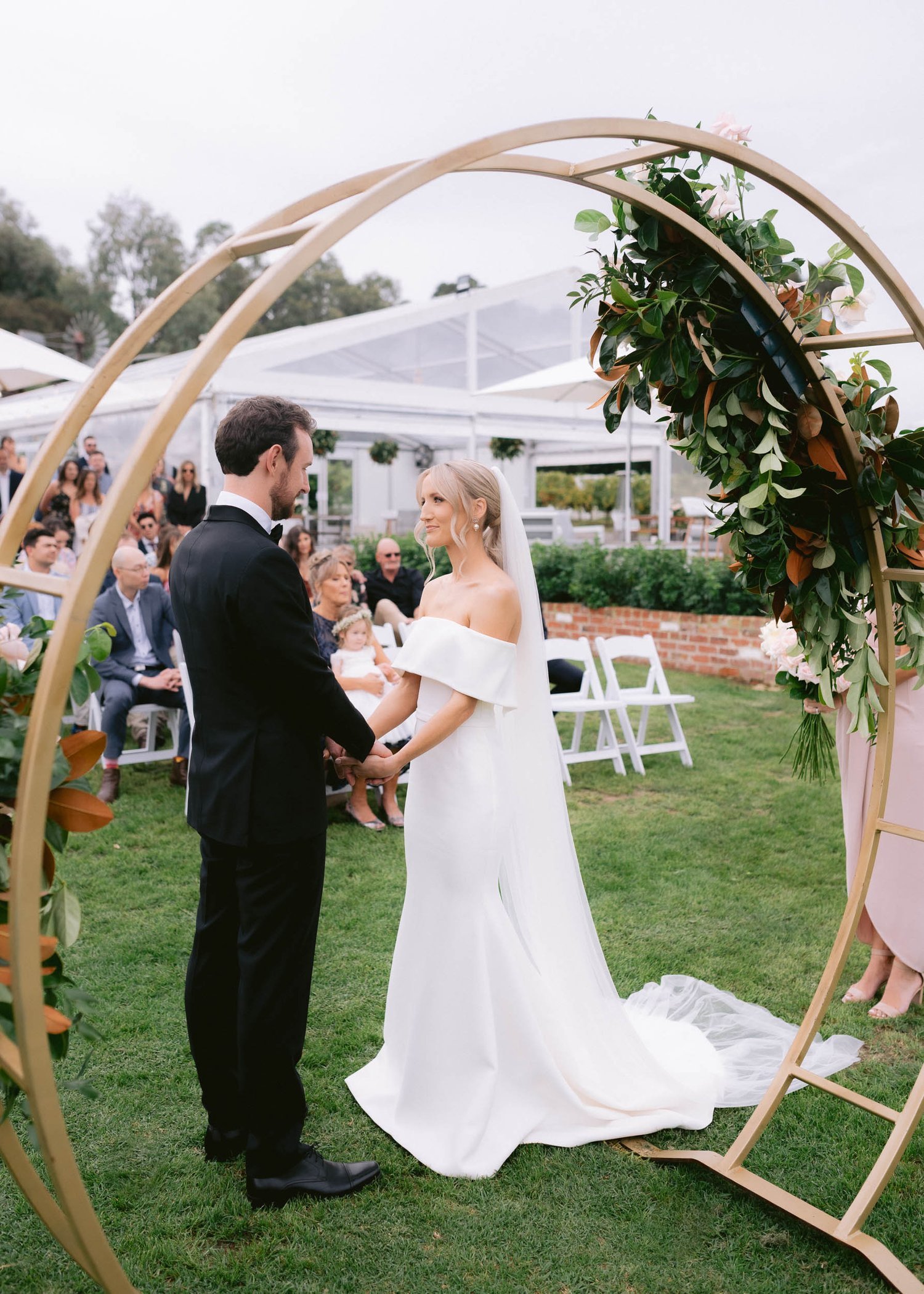A bride and groom holding hands during an outdoor wedding ceremony, under a decorative arch adorned with greenery. Guests are seated in the background on white chairs. The bride is wearing a white off-the-shoulder gown, and the groom is in a black su