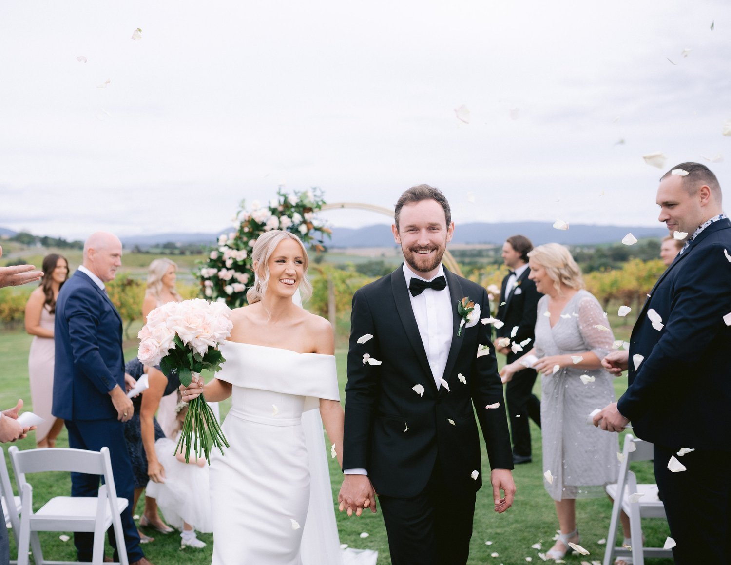 Bride and groom walking down the aisle, smiling, with guests in the background. Bride holding a bouquet of pink roses. Wedding ceremony outdoors with rose petal confetti.