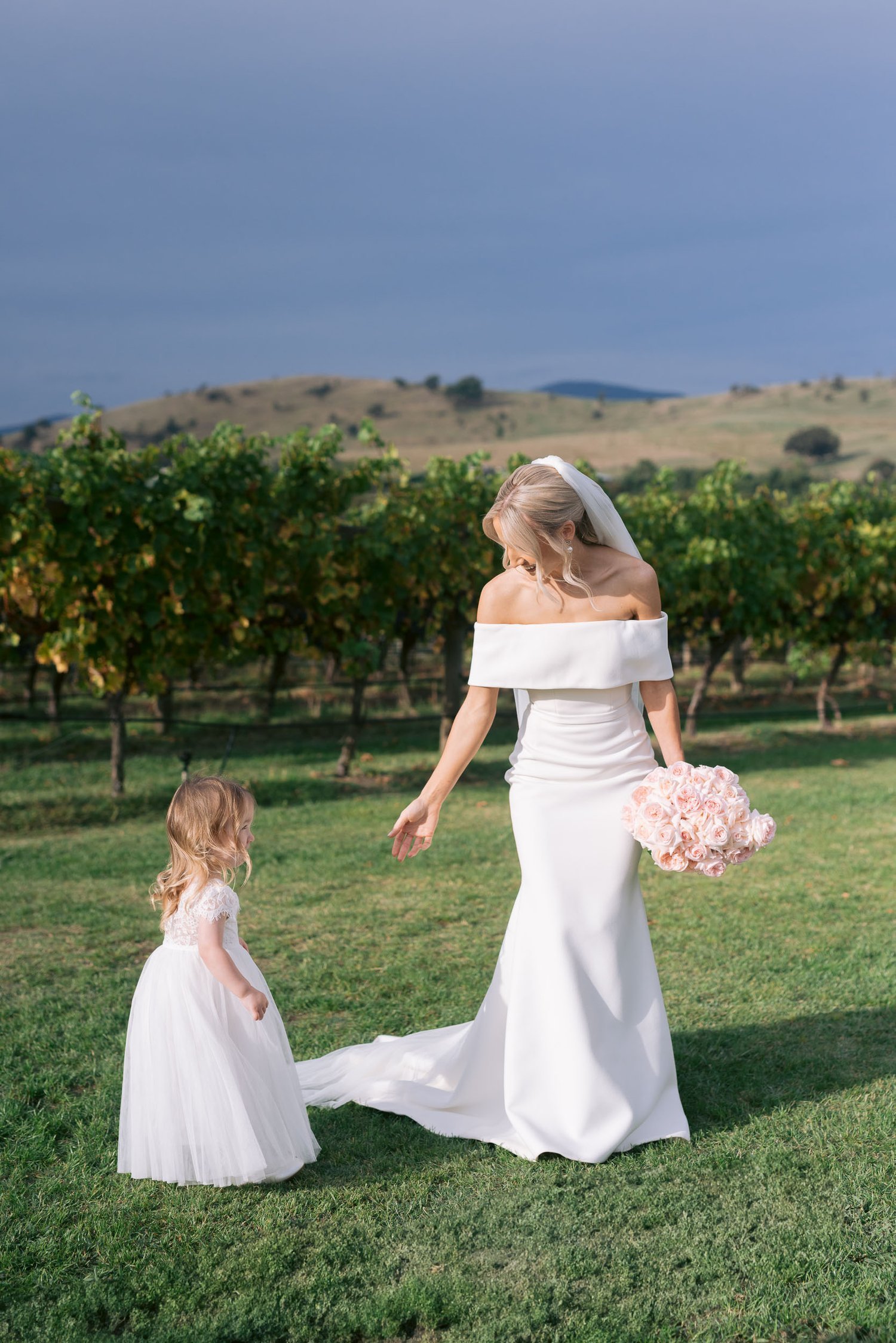 Bride in a white dress with a bouquet of flowers and a flower girl in a vineyard setting.