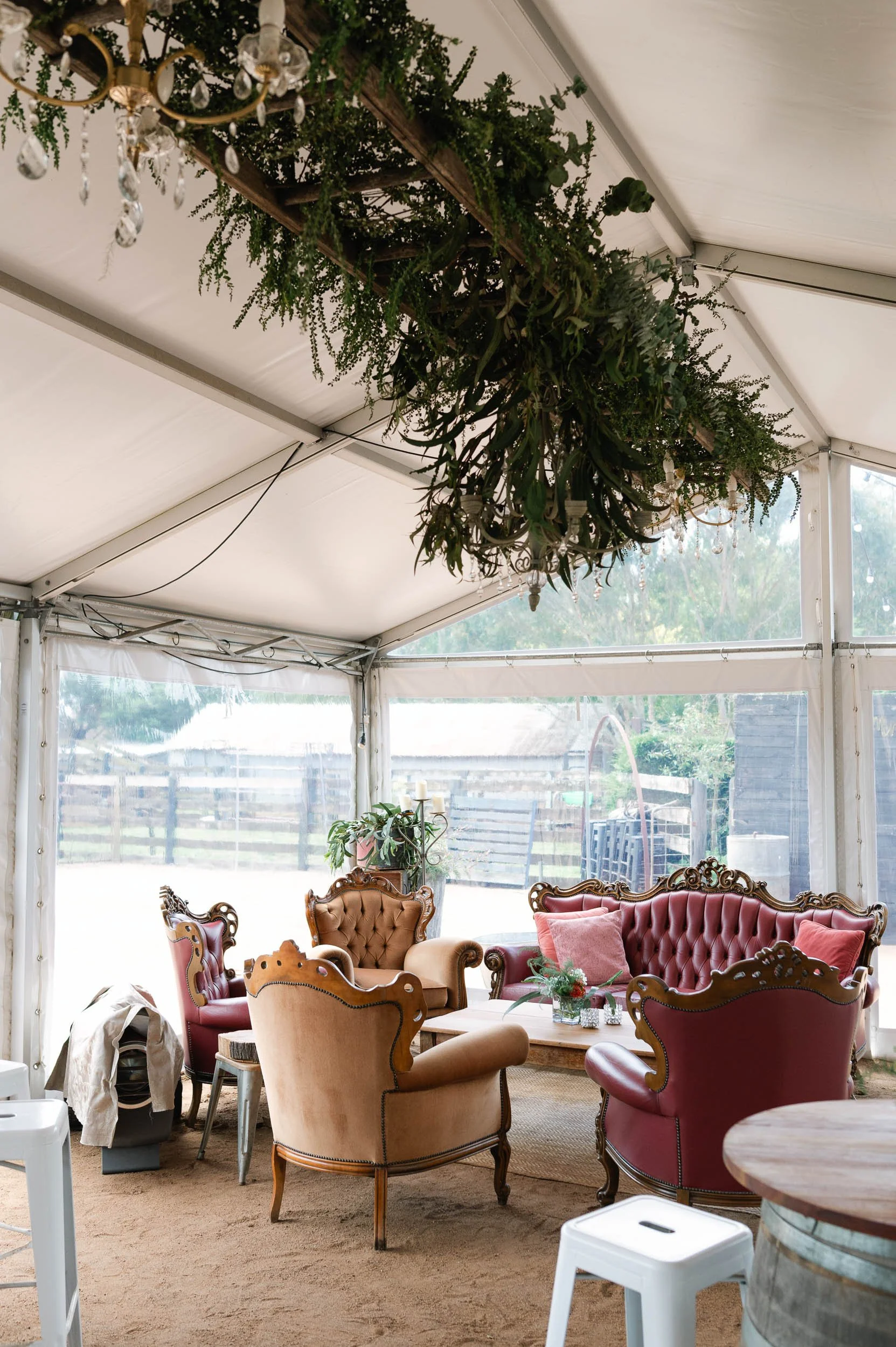 Vintage-style outdoor seating area inside a tent with tufted sofas, armchairs, a hanging chandelier, and a lush green foliage overhead.