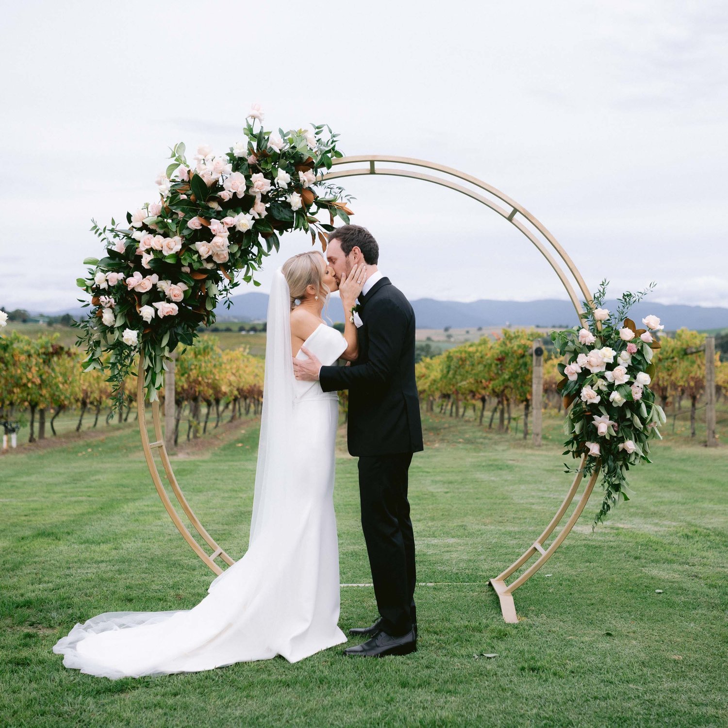 Bride and groom kissing under a floral arch in an outdoor vineyard wedding ceremony.