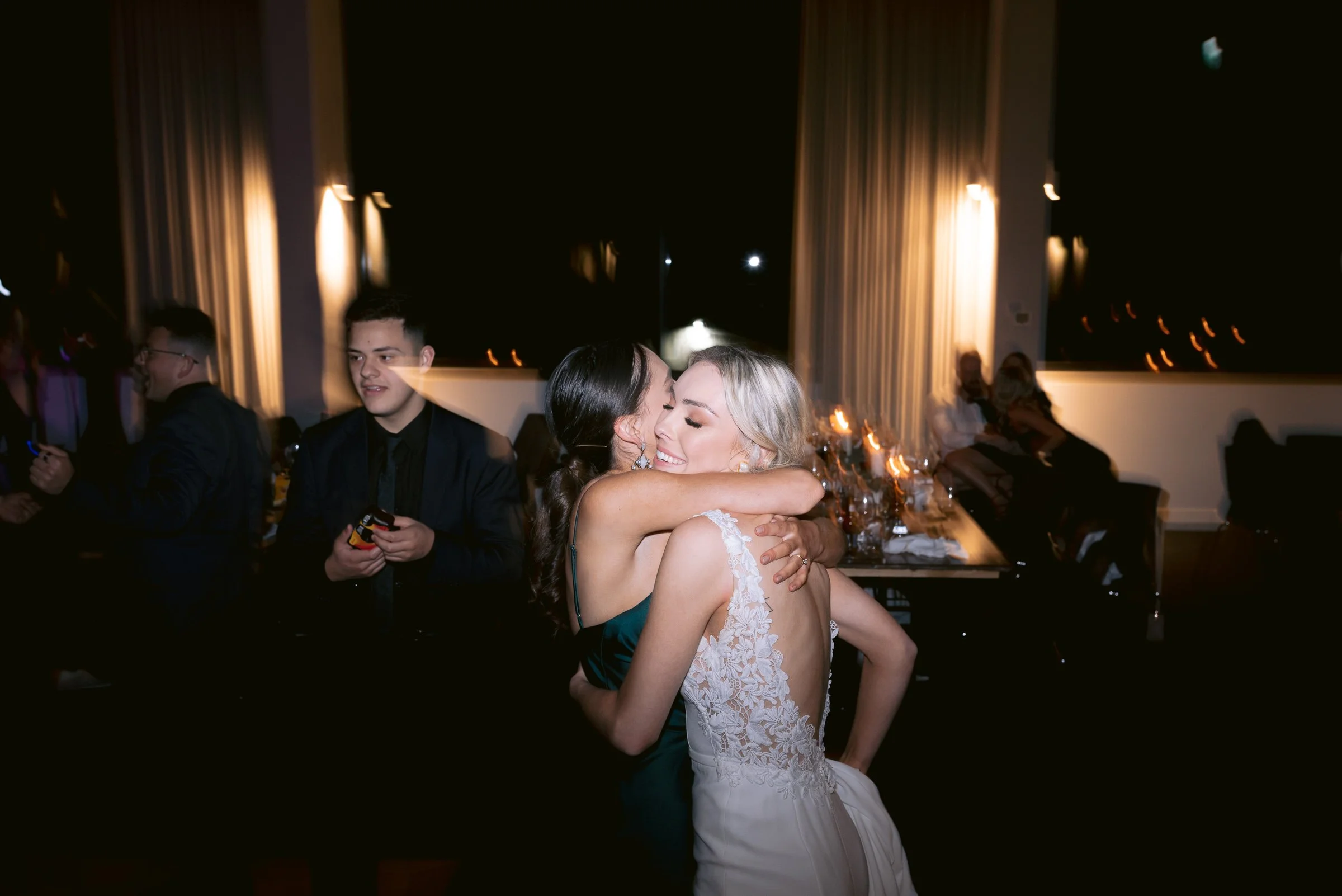 Two women hugging at an indoor event, with people and tables visible in the background.