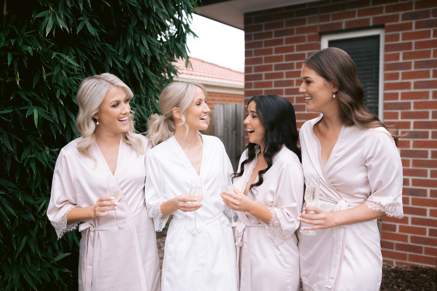 Four women in silk robes holding glasses of champagne, standing outdoors by a brick wall and greenery, smiling and talking.