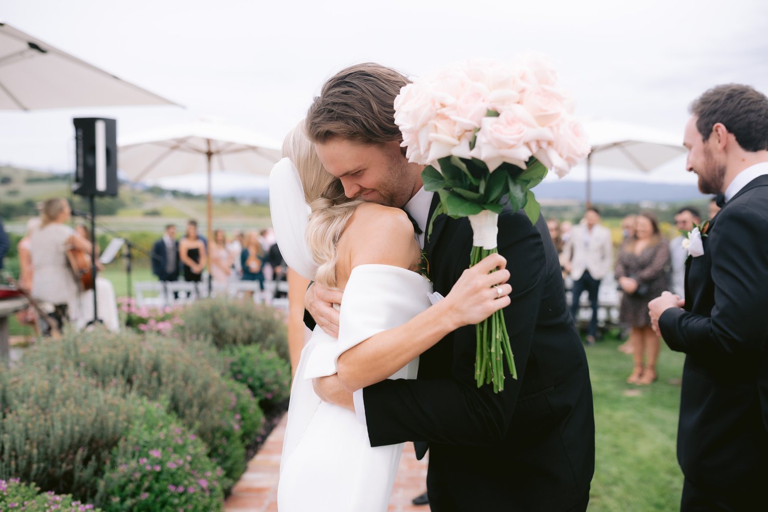 Bride and groom hugging outdoors, holding pink roses, surrounded by guests at a wedding ceremony.