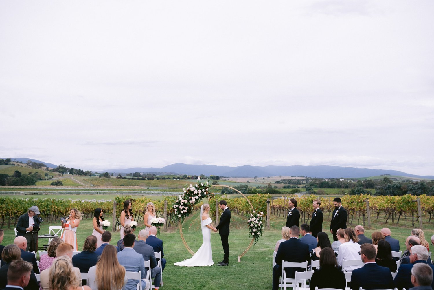 Outdoor wedding ceremony in a vineyard with a circular floral arch; bride and groom exchanging vows, surrounded by bridesmaids and groomsmen. Guests seated in the foreground.
