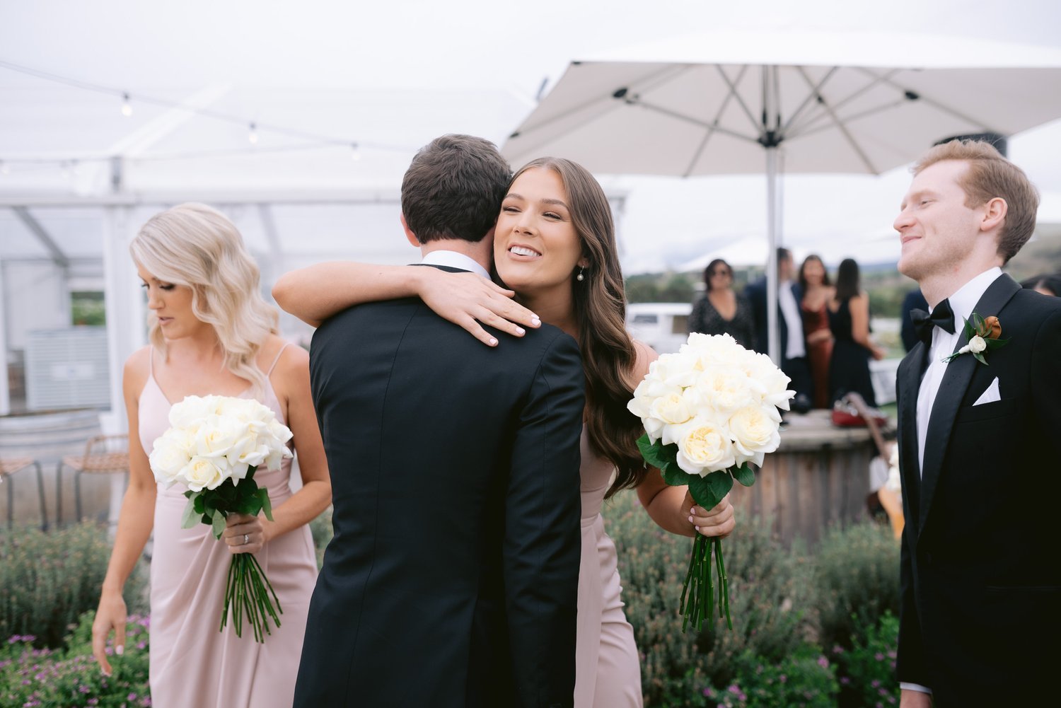 Wedding scene with bridesmaids holding white rose bouquets, a man in a suit being hugged, and another man smiling, outdoor setting, white umbrellas in background.
