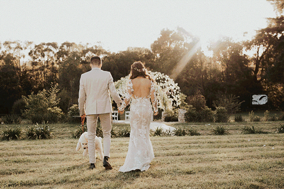 Bride and groom walking hand in hand outdoors, accompanied by a dog, towards a floral wedding arch, in a sunlit garden setting.