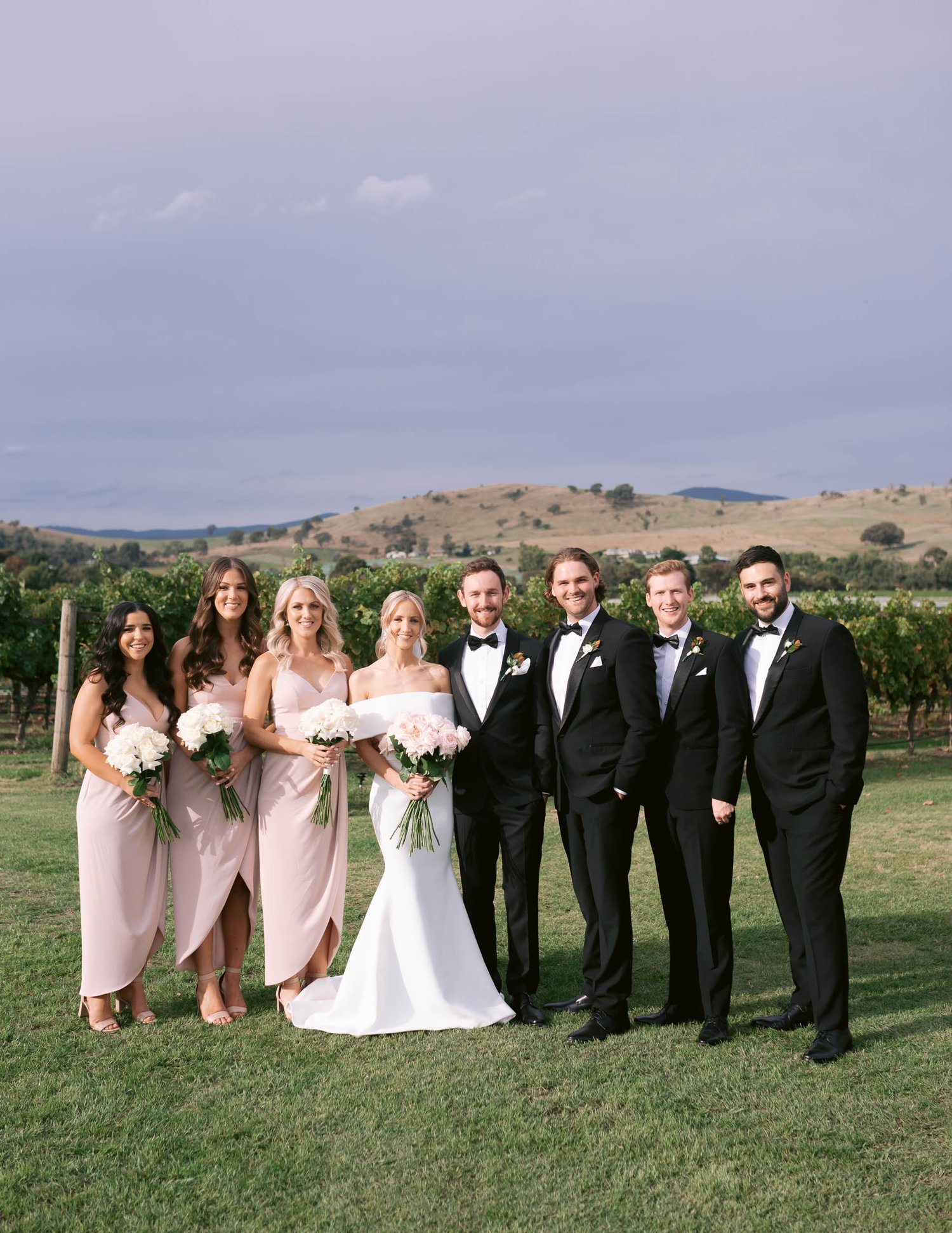 Wedding party posing outdoors with groomsmen in black suits and bridesmaids in pink dresses holding flowers, set in a vineyard with hillside background at Acacia Ridge Winery, Yara Valley.