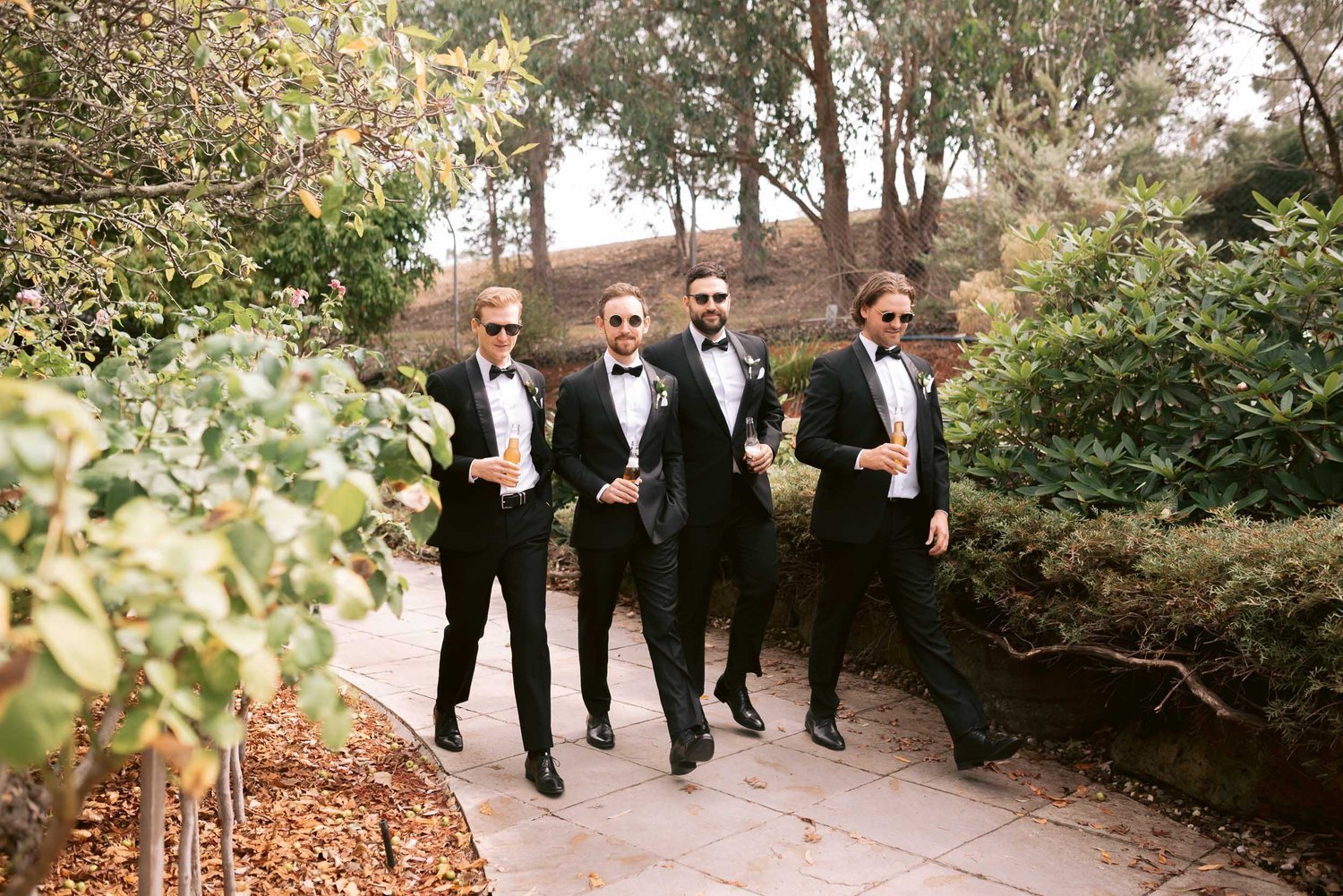 Four men in tuxedos walking on a garden path, holding bottled drinks.