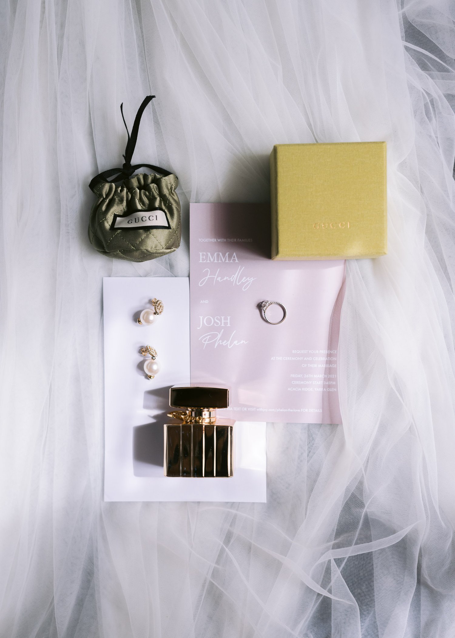 Flat lay of wedding invitation, engagement ring, Gucci jewelry pouch, yellow Gucci gift box, pearl earrings, and a perfume bottle on a white tulle fabric background.