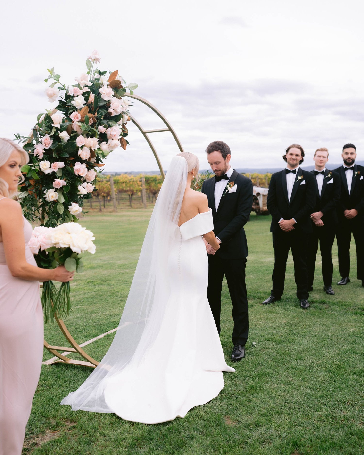 Outdoor wedding ceremony with bride in white dress and groom in black suit standing by floral arch, bridesmaid holding bouquet, and groomsmen in the background.