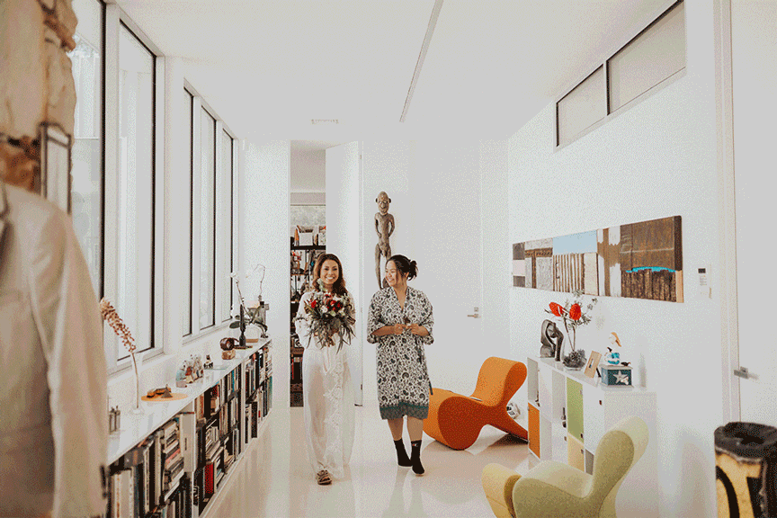 Two women walking through a modern hallway with large windows, bookshelves filled with books, and art pieces on the walls.