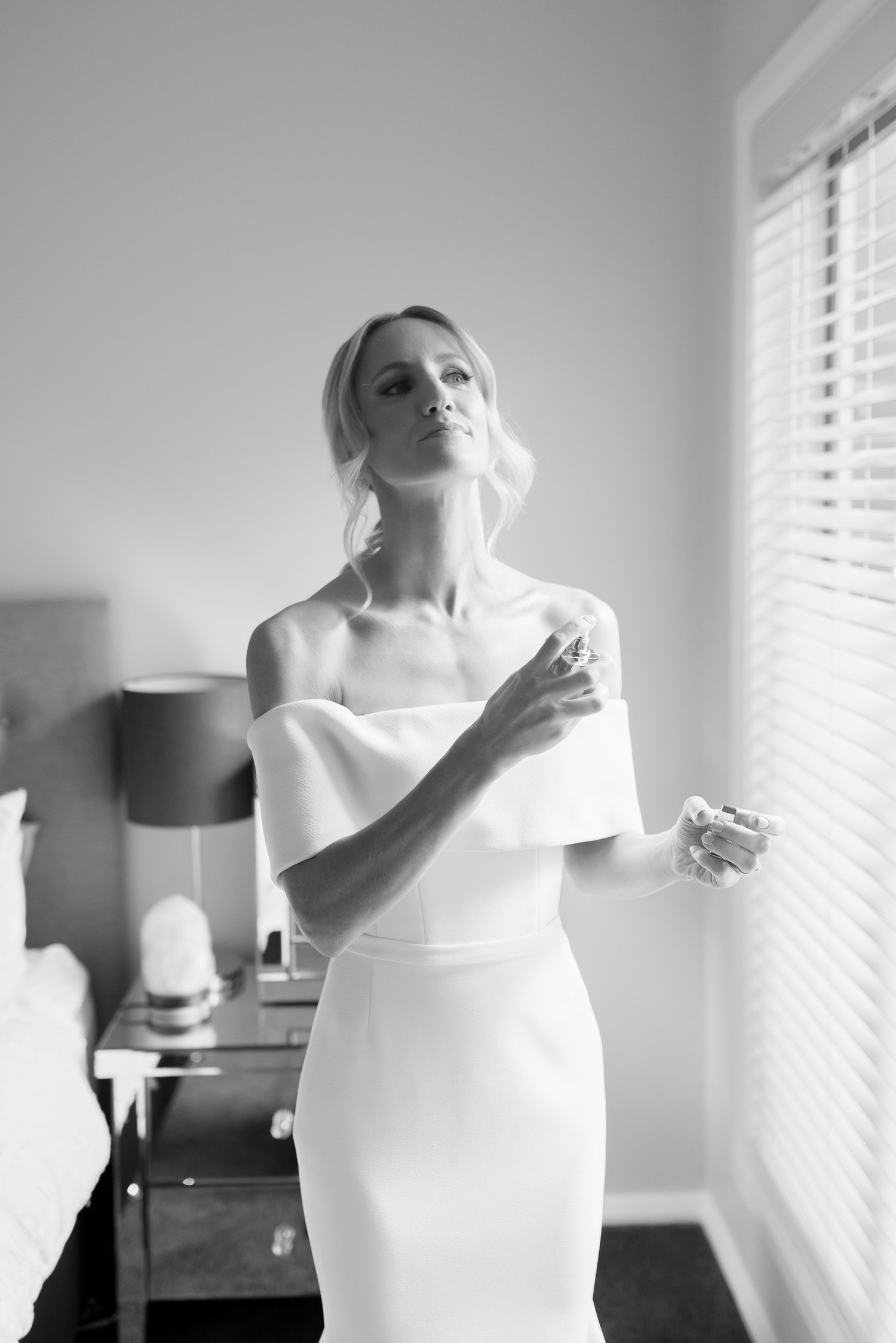 Black and white photo of a woman in a white off-the-shoulder dress applying perfume in a bedroom with a lamp and side table.