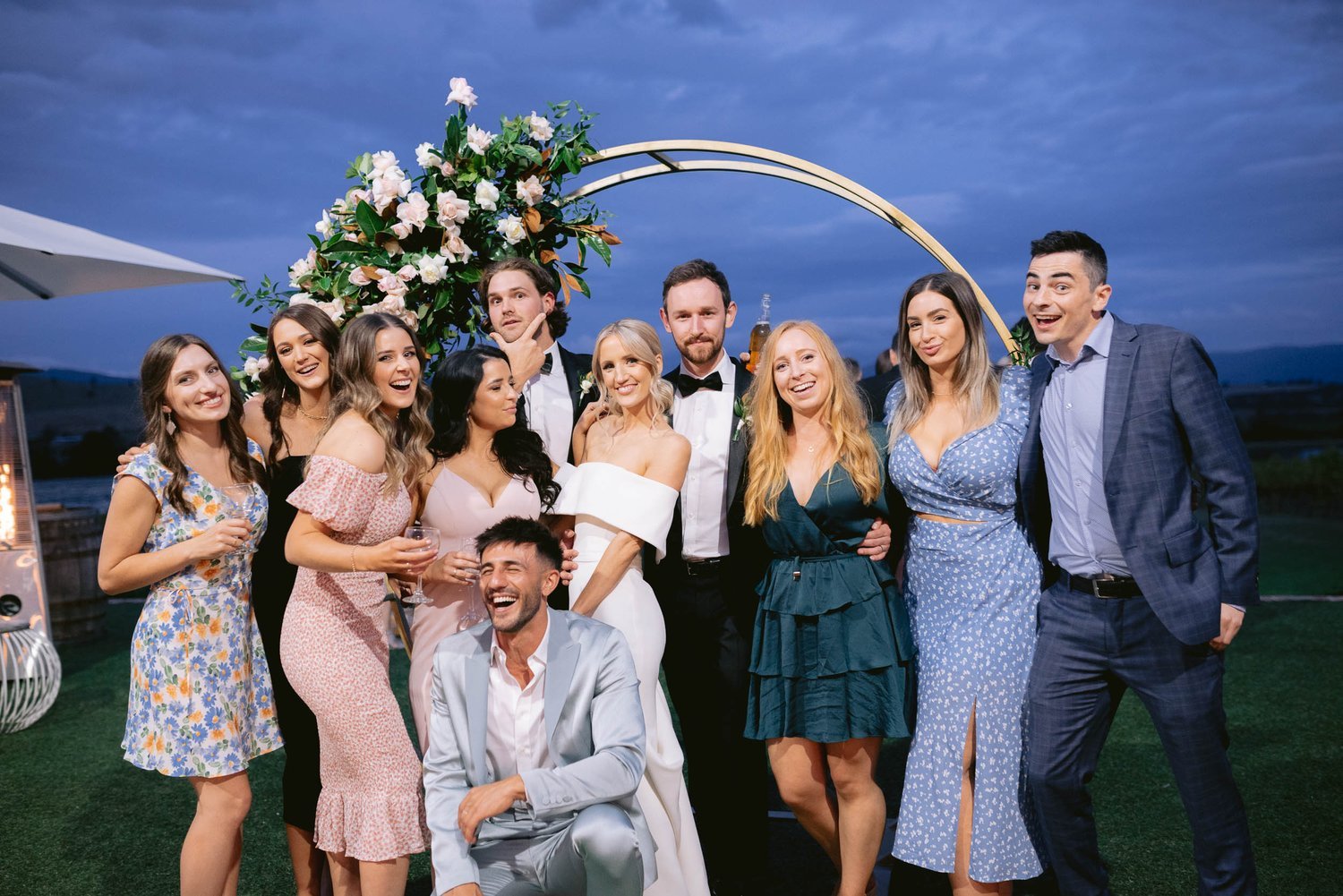 Group of people smiling and posing at an outdoor wedding event, standing in front of a floral archway, with a blue evening sky backdrop.