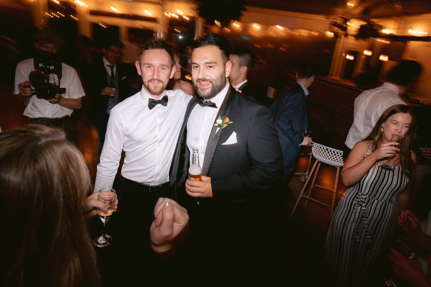 Two men in formal attire posing at a party, one holding a beer. Guests and a photographer in the background. Woman drinking on the right.
