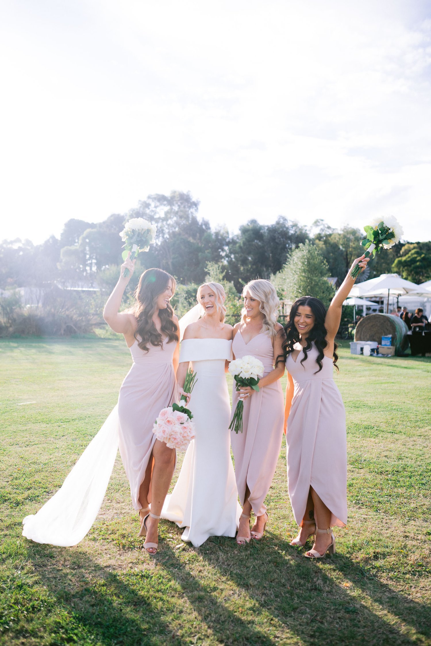 Bride with three bridesmaids in pastel dresses holding bouquets, smiling outdoors on a sunny day.