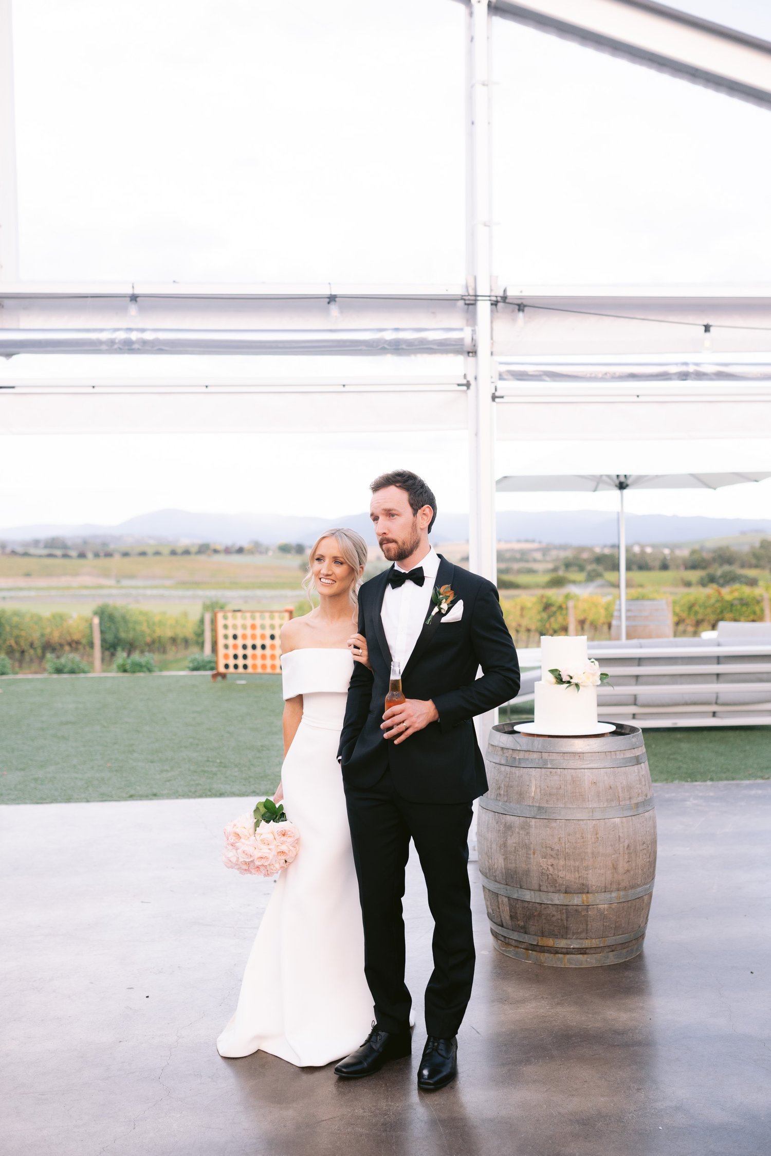 A bride and groom stand together at an outdoor wedding venue with mountains in the background. The bride wears a white gown and holds a bouquet of pink roses. The groom is in a black tuxedo, holding a beer bottle. A cake sits on a barrel to their sid