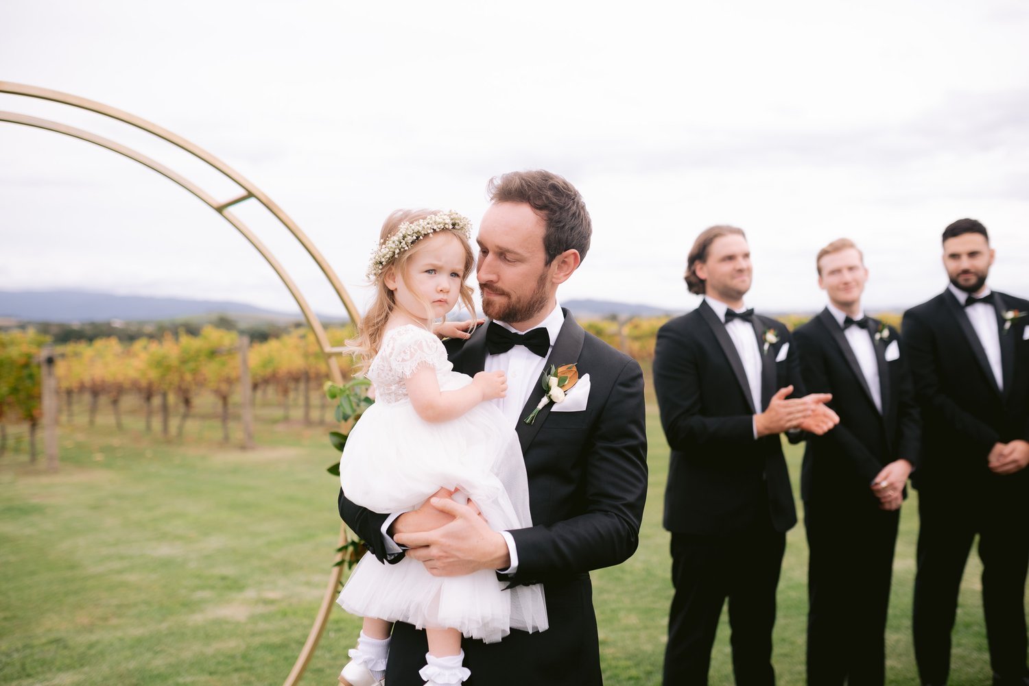 A wedding scene with a man in a tuxedo holding a young girl in a white dress and floral headband. They stand outdoors, with a group of men in tuxedos clapping in the background, and a vineyard visible in the distance.