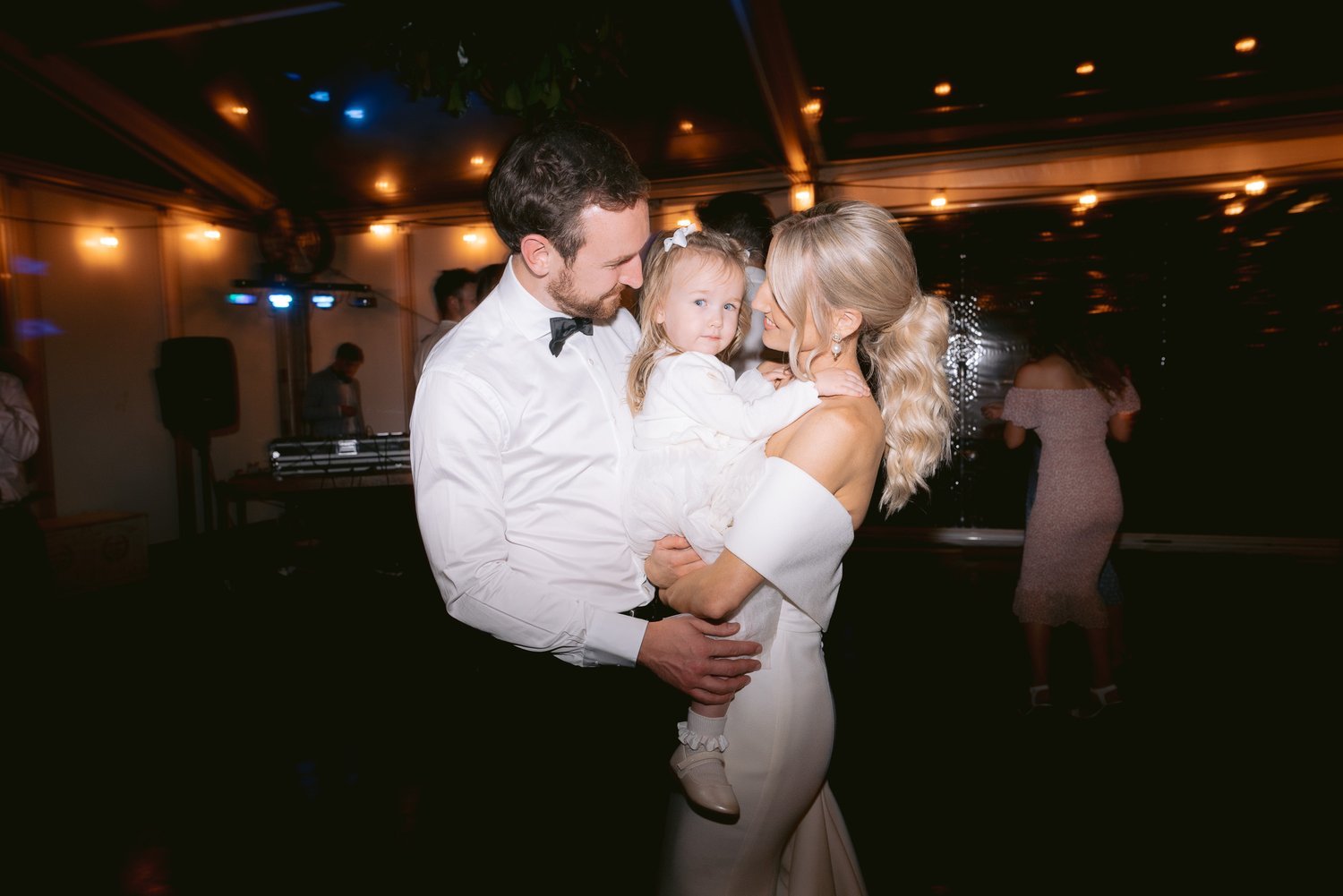 A couple in formal attire, with the man wearing a white shirt and bow tie and the woman in a white dress, holding a child in a dimly lit venue with string lights.