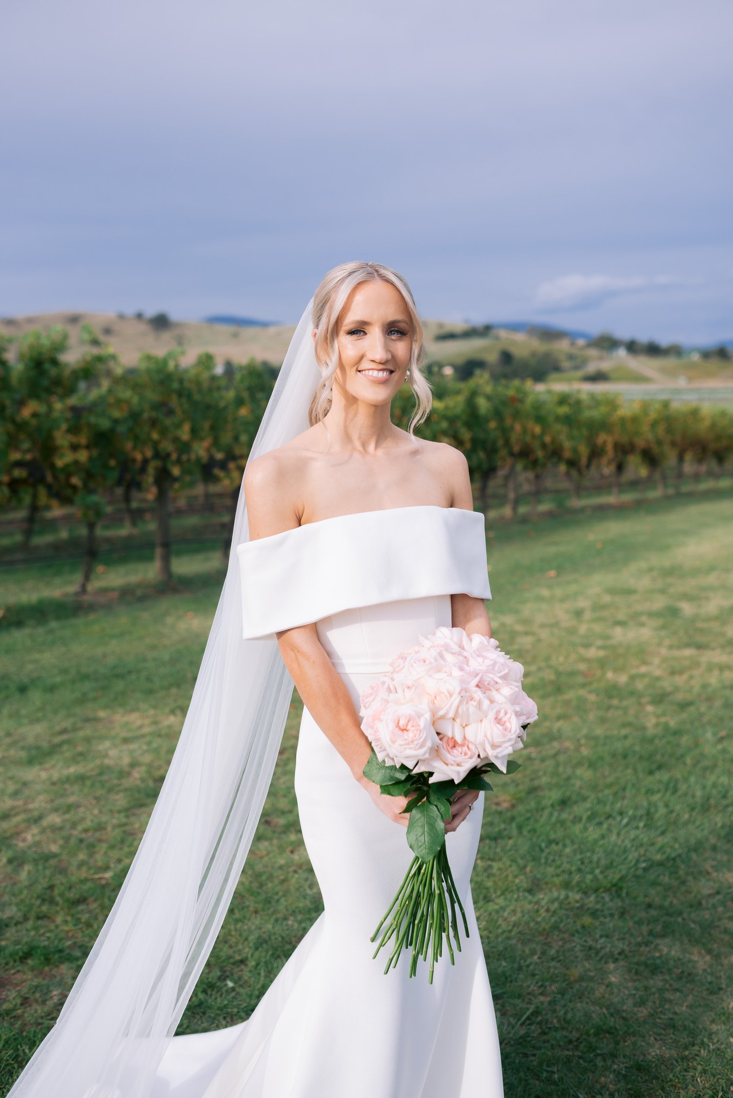 Bride in an off-the-shoulder white wedding dress holding a bouquet of pink flowers, standing in a vineyard setting.