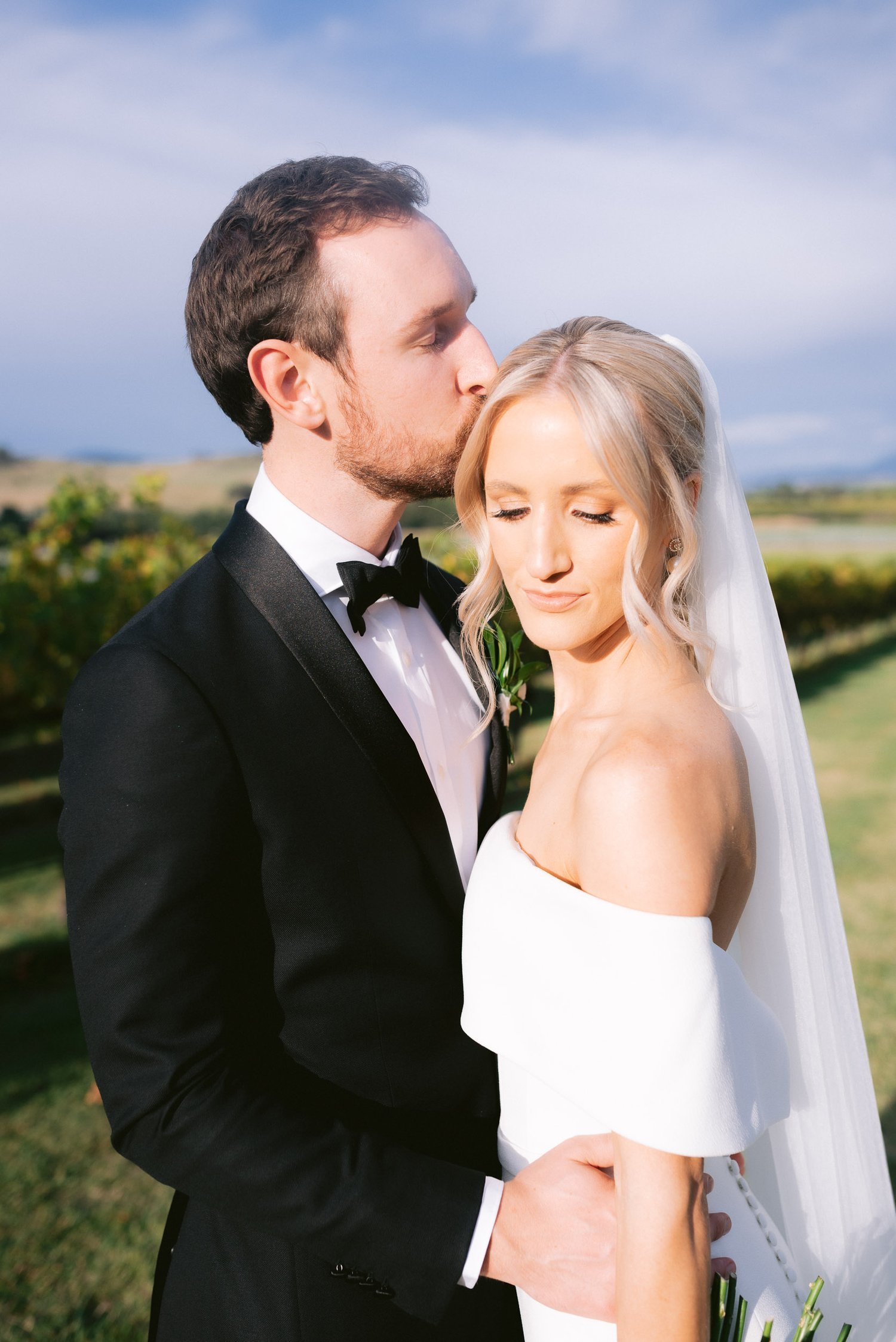 Bride and groom posing outdoors, groom kissing bride's forehead. Bride in white off-shoulder dress with veil, groom in black tuxedo.