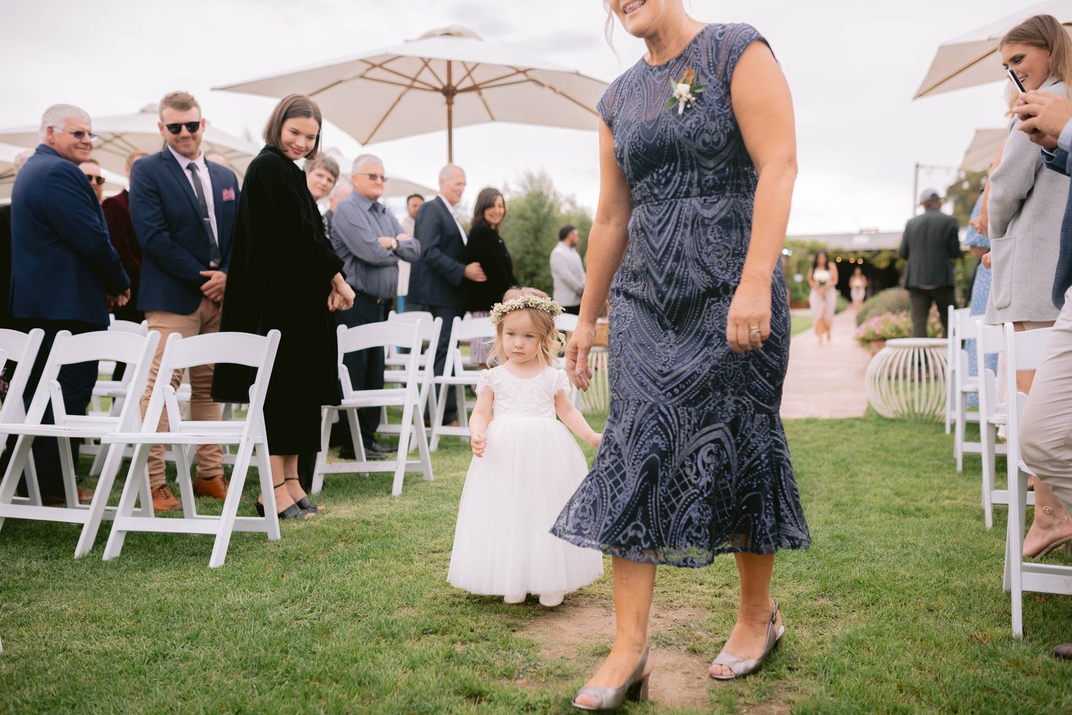 A wedding scene with a flower girl in a white dress walking down an aisle, holding hands with a woman in a blue patterned dress, surrounded by seated and standing guests.