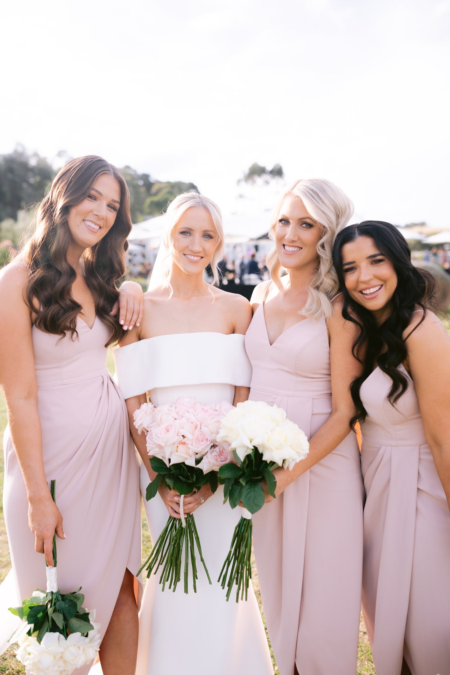 Bride and bridesmaids in pastel dresses holding bouquets, smiling outdoors at a wedding.
