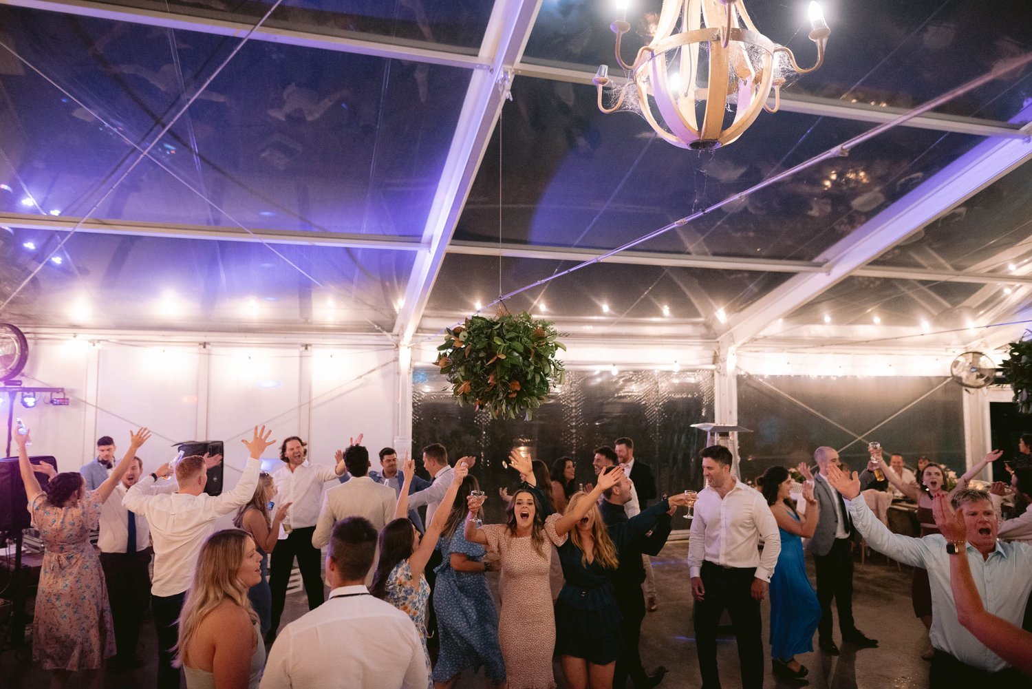 People dancing and celebrating at a party under a large transparent tent with string lights, chandelier, and greenery decoration in the Yarra Valley.