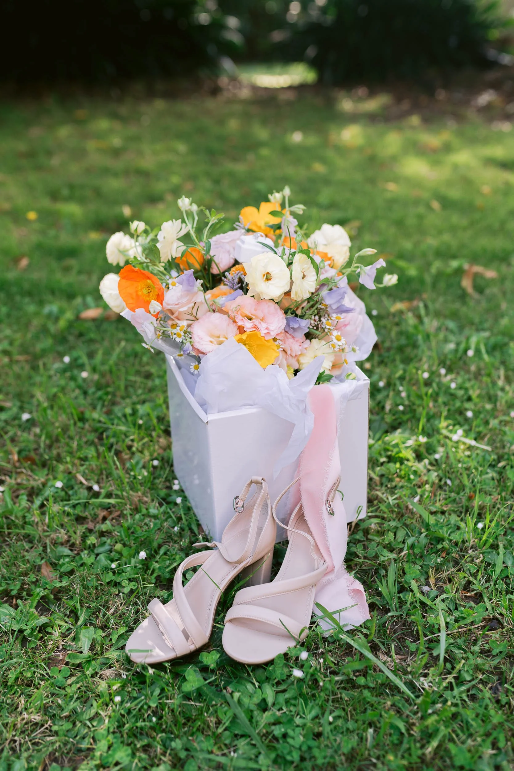 A bouquet of colorful flowers in a white box with a pair of beige high-heeled sandals on grass.
