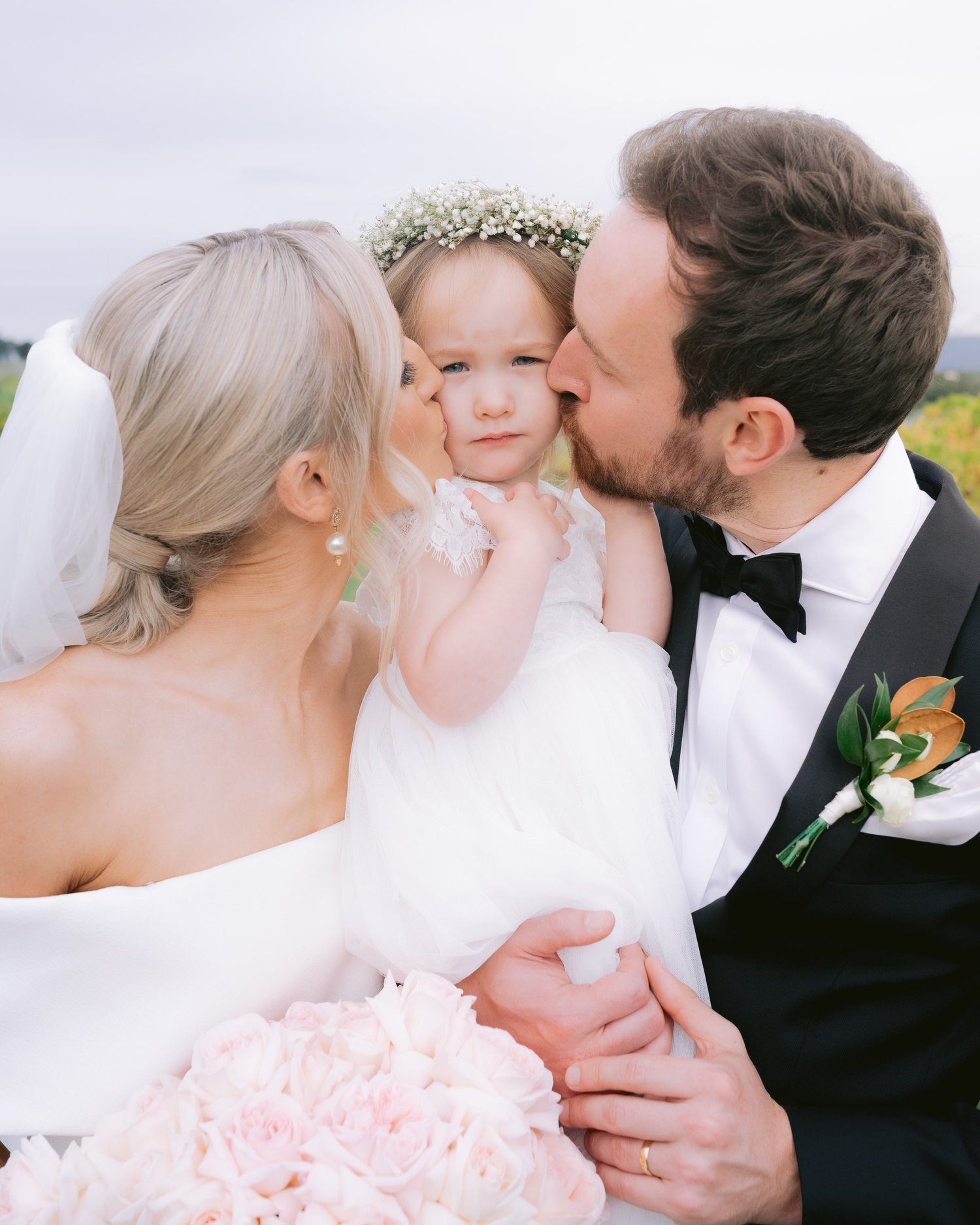 Bride and groom kissing a child in a white dress at a wedding ceremony.