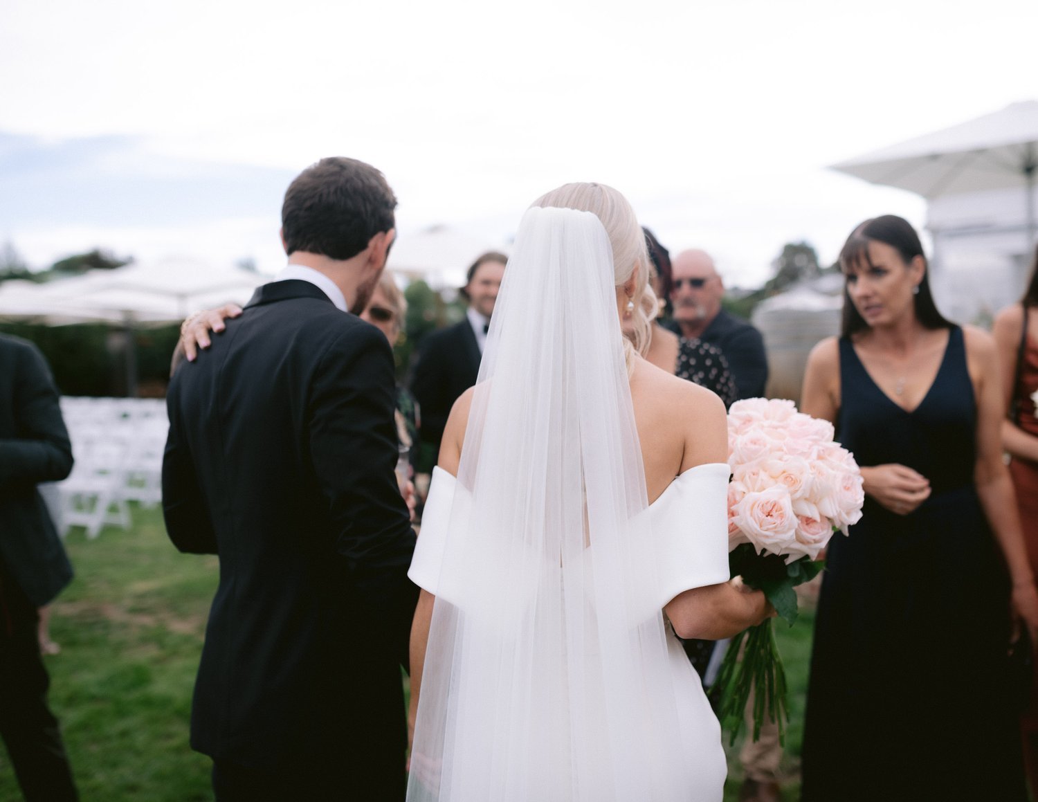 Bride and groom at an outdoor wedding ceremony, with the bride holding a bouquet of pink roses and wearing a veil.