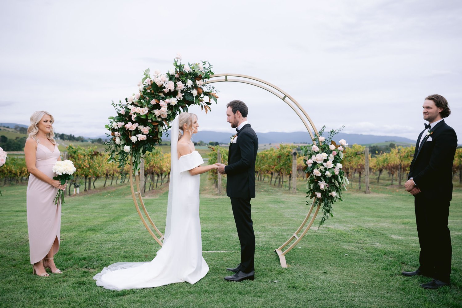 Outdoor wedding ceremony with a couple exchanging vows under a floral arch, attended by a bridesmaid and groomsman, set in a vineyard.
