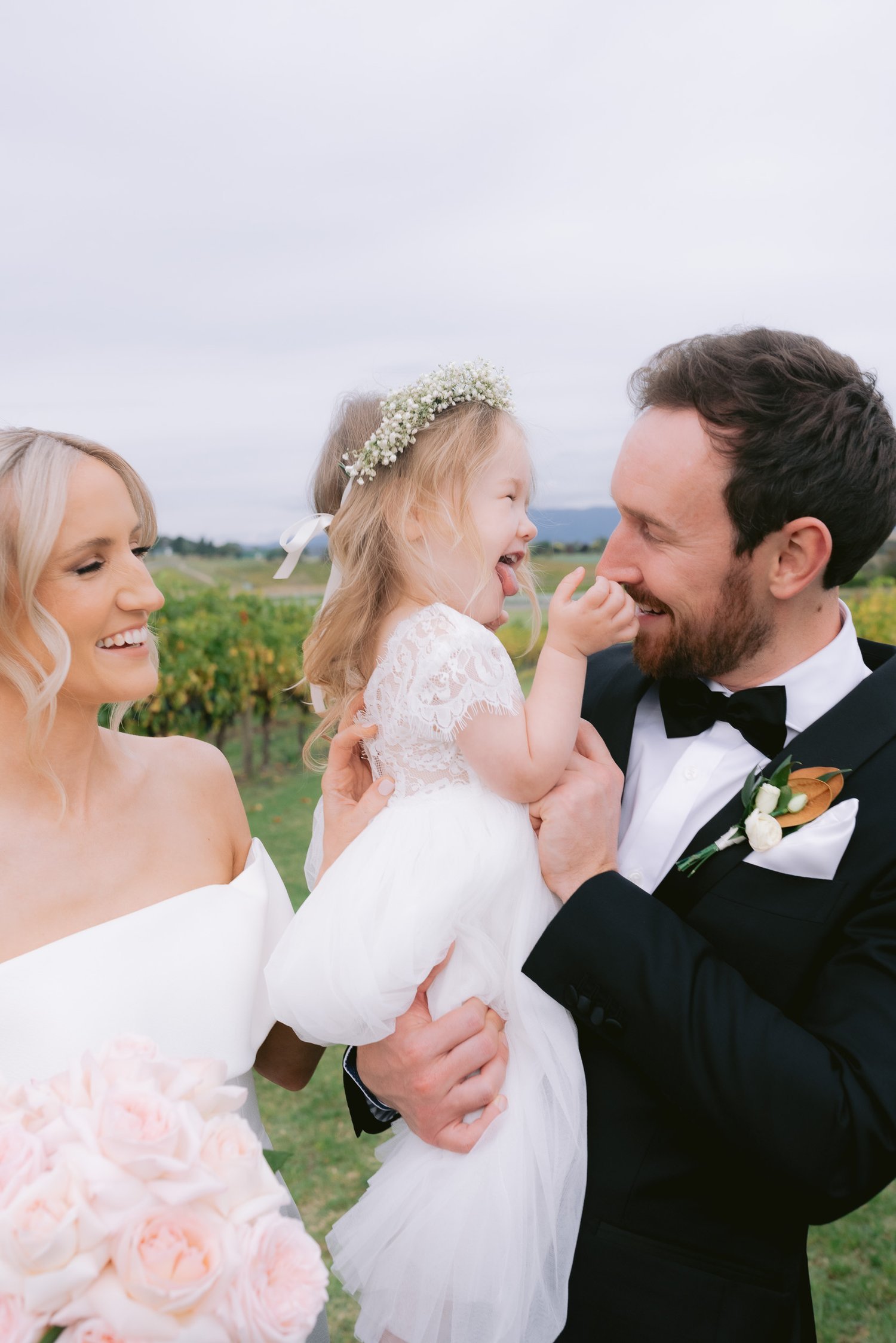 Bride and groom smiling at a young girl in a white dress and flower crown at an outdoor wedding.