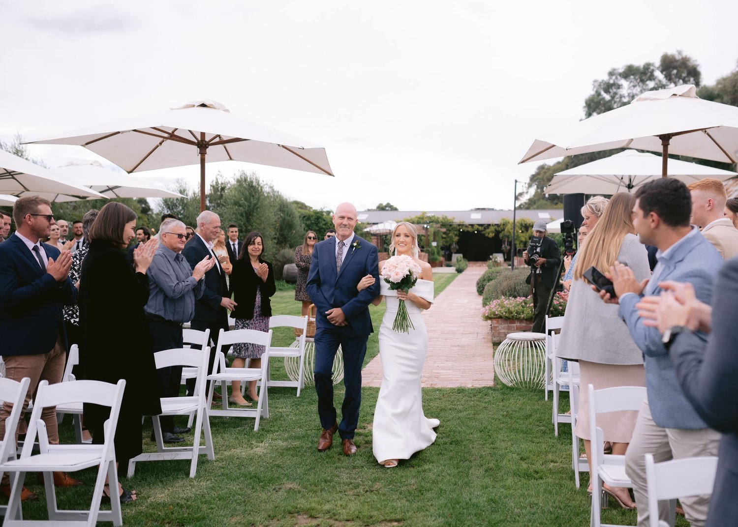 Bride walking down the aisle with a man in a garden wedding setting, surrounded by applauding guests seated on white chairs under large umbrellas.