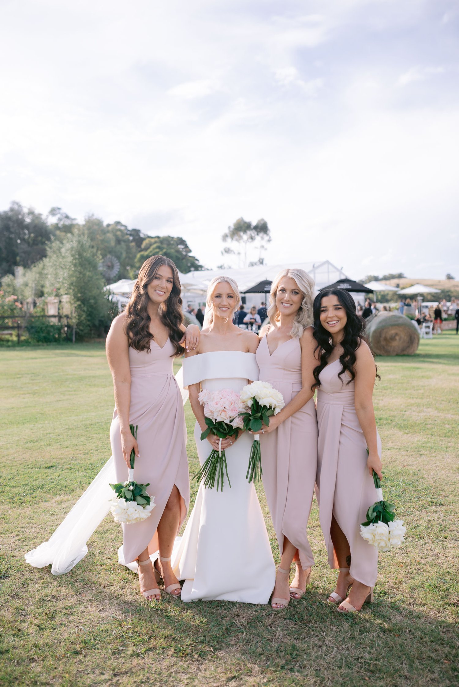 Bride and bridesmaids posing outdoors in a grassy area, wearing pink and white dresses, holding bouquets, with trees and outdoor tents in the background.