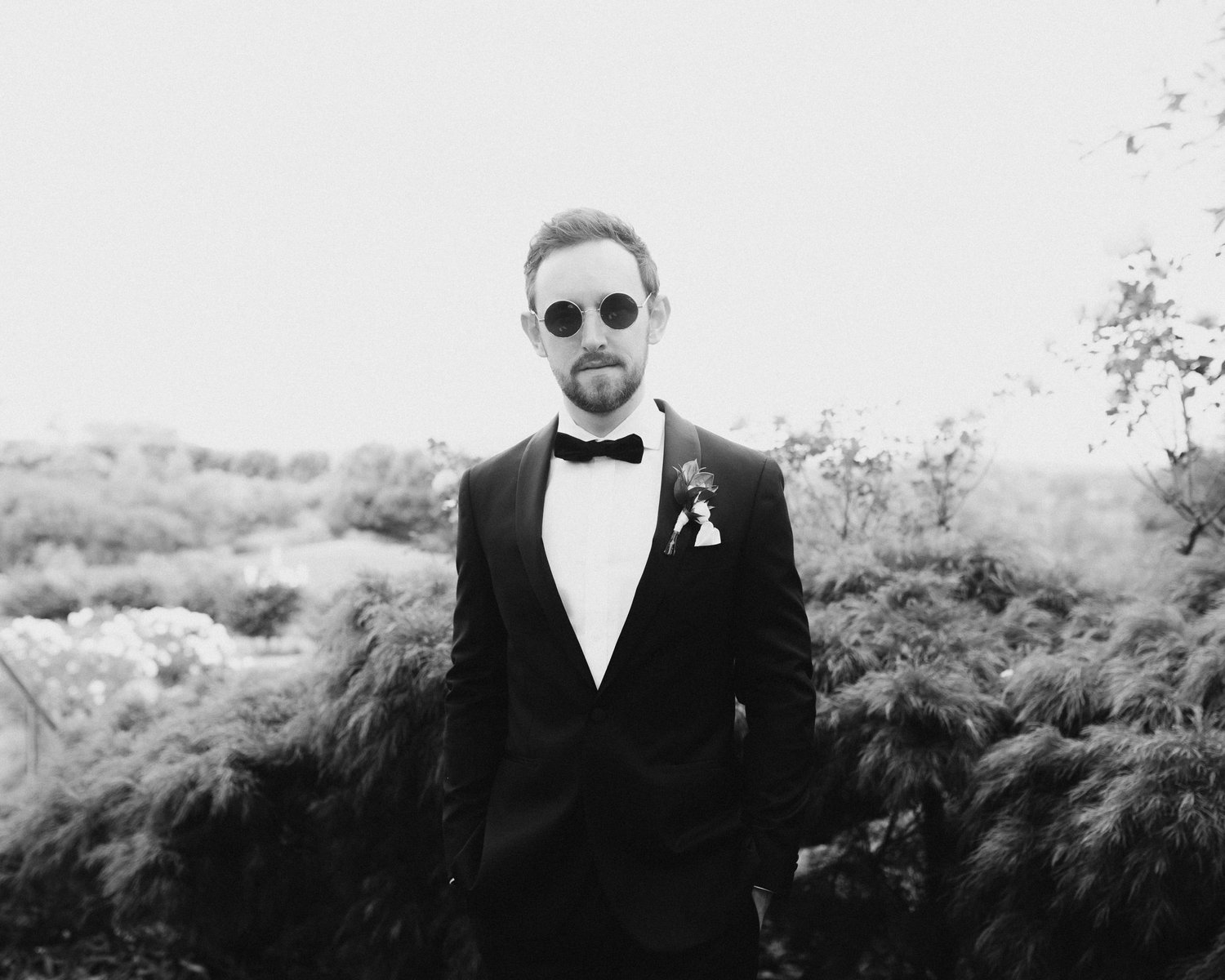 Black and white photo of a man in a tuxedo with sunglasses, standing outdoors with trees in the background.