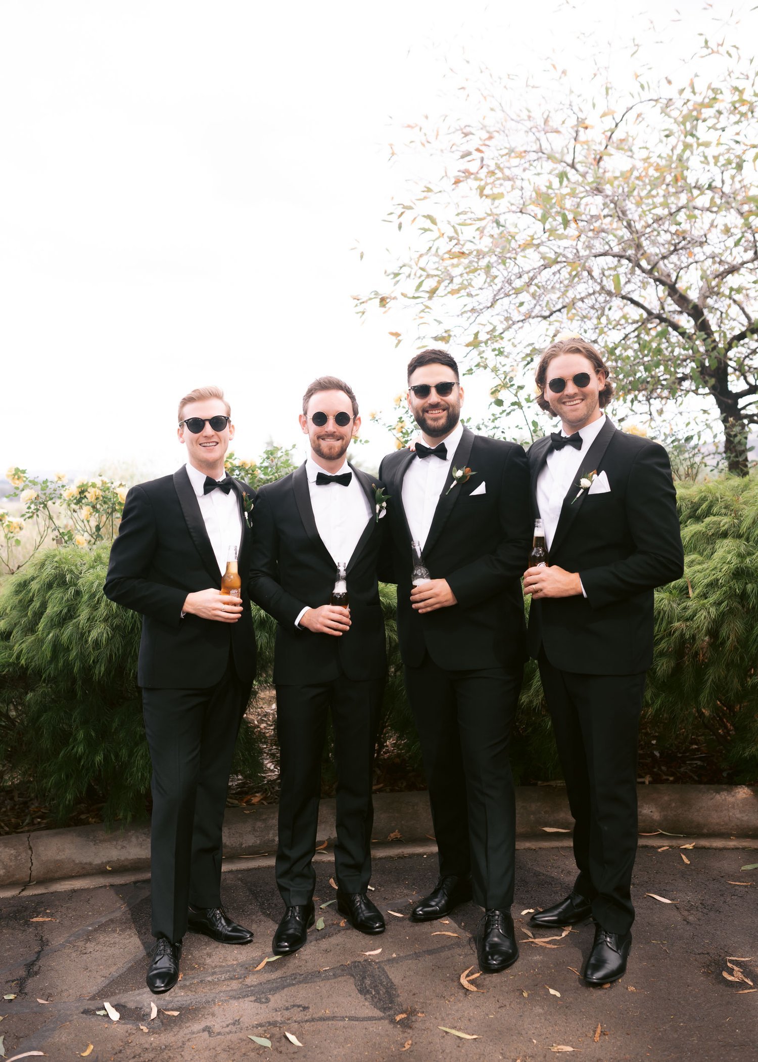Four men wearing black tuxedos and sunglasses, standing outdoors, holding bottles, with trees and greenery in the background.